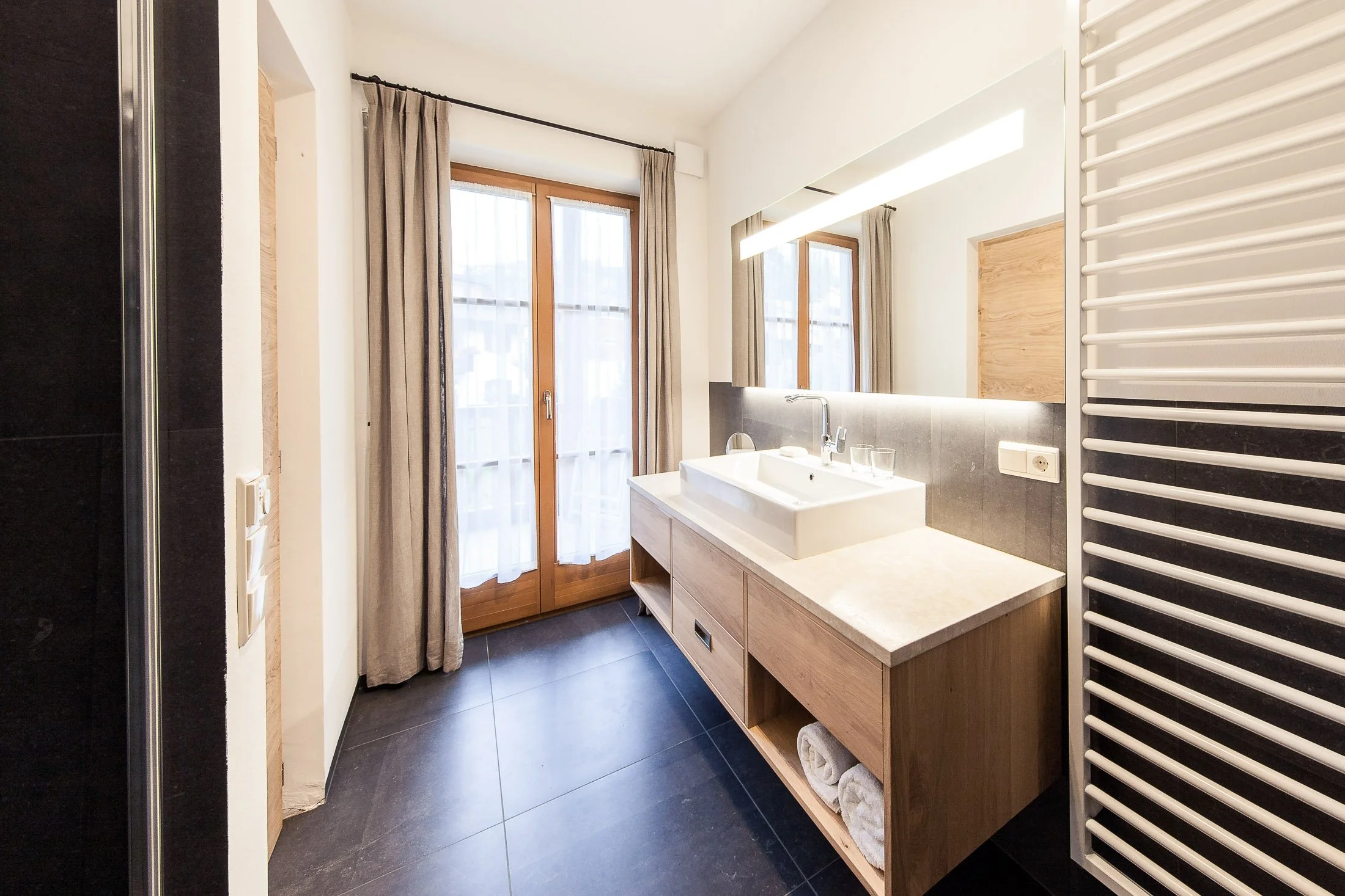 Badezimmer mit Waschbecken, großen Fenstern, grauen Fliesen, Handtücher und einem Heizkörper