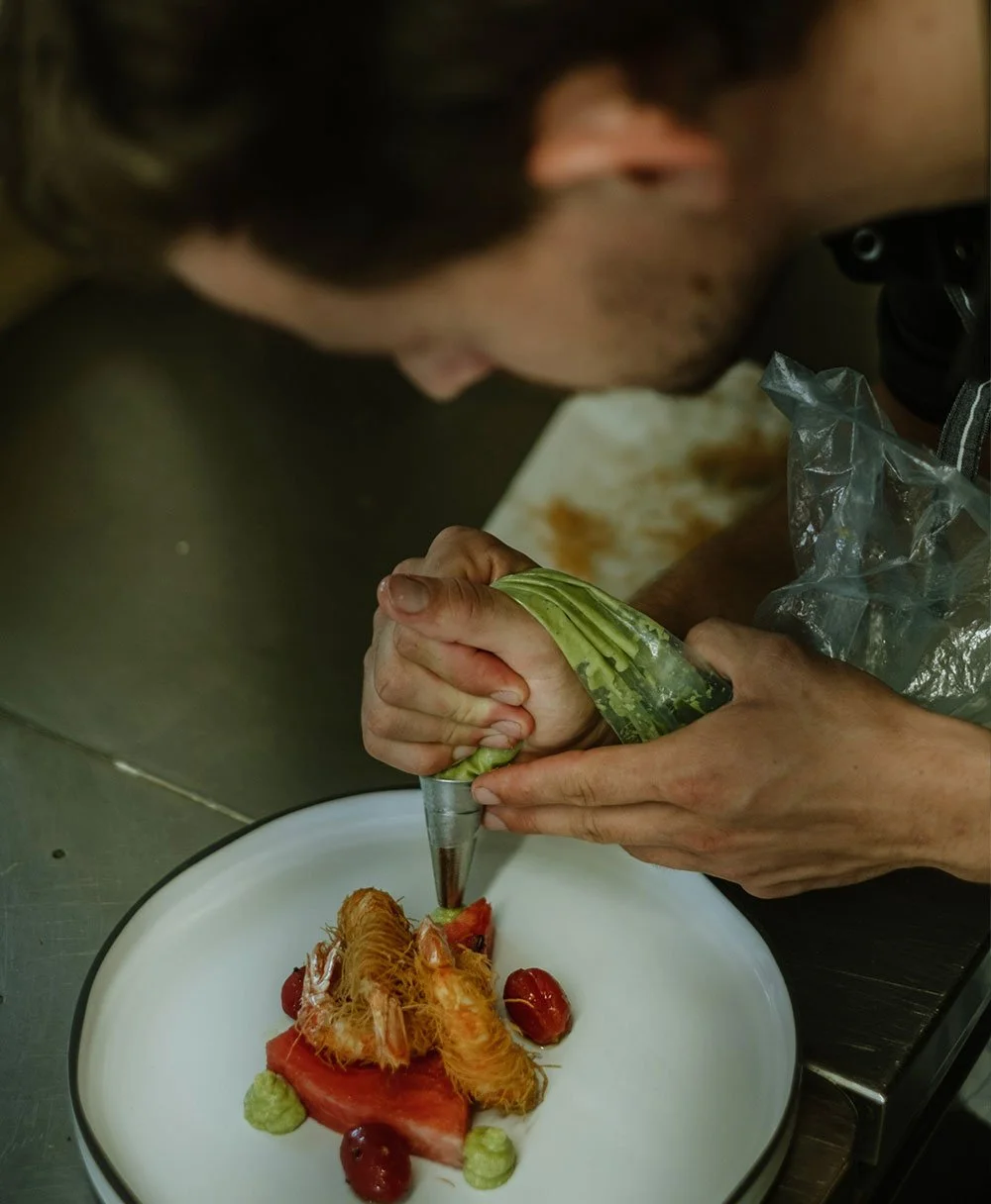 Ein Mann gestaltet ein Gericht mit einer Spritzpistole, das zeigt Garnelen, Tomate, Gurke und Beeren auf einem weißen Teller, in einem professionellen Küchenumfeld.