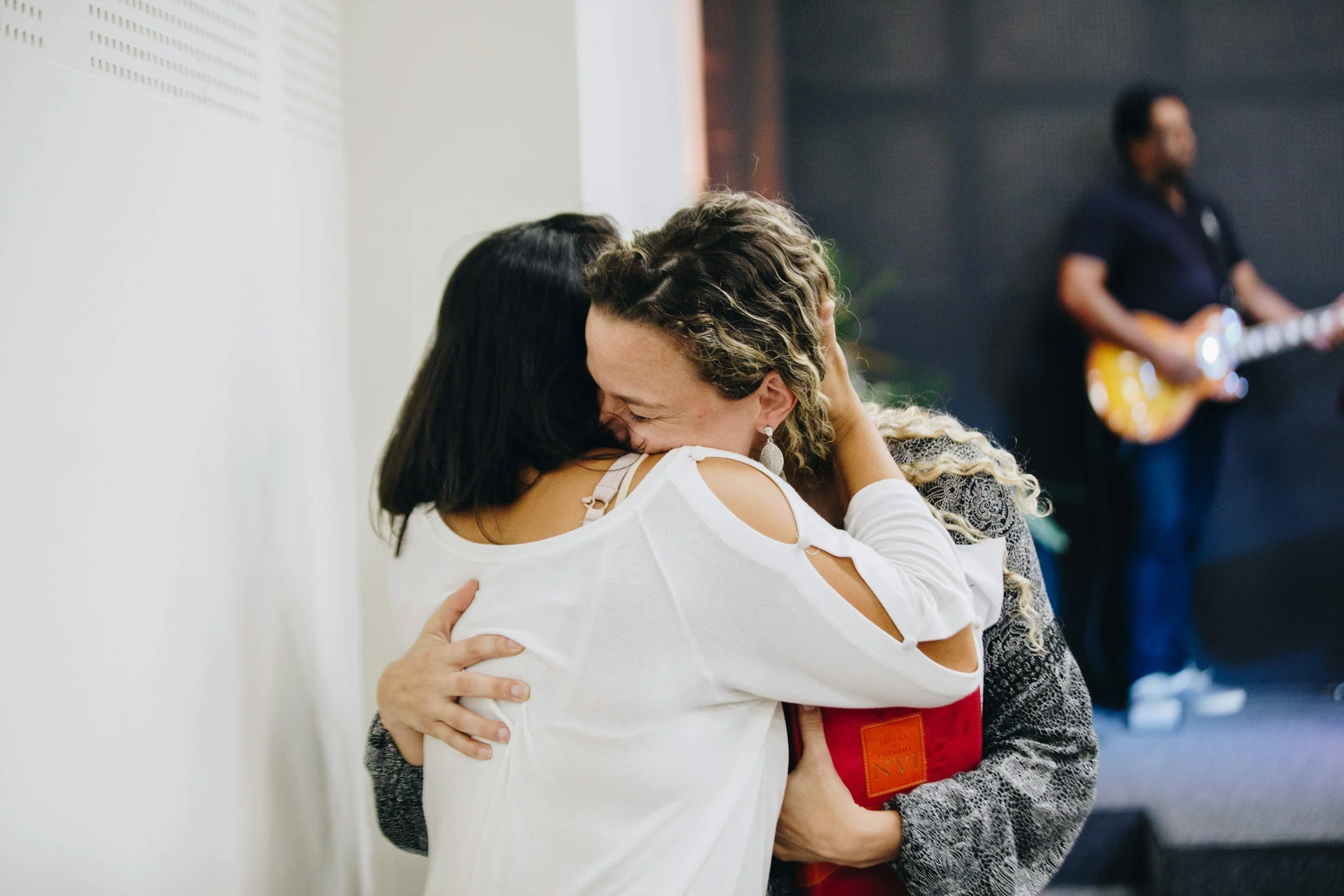 Two women hugging warmly in an indoor setting with a person playing electric guitar in the background.