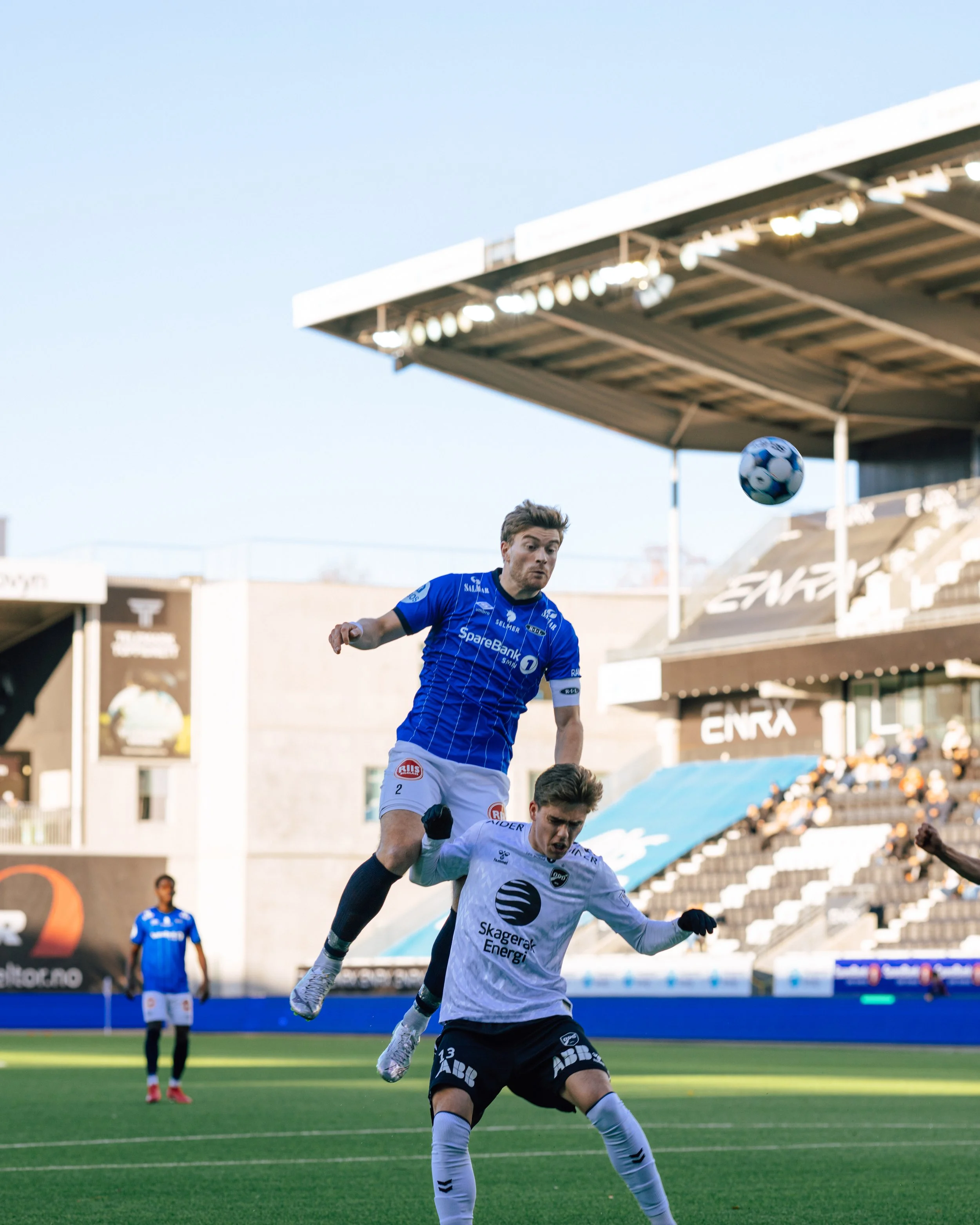 Two soccer players jump to head the ball during a match. One player in a blue jersey is in mid-air, while the other in a white jersey is on the ground.