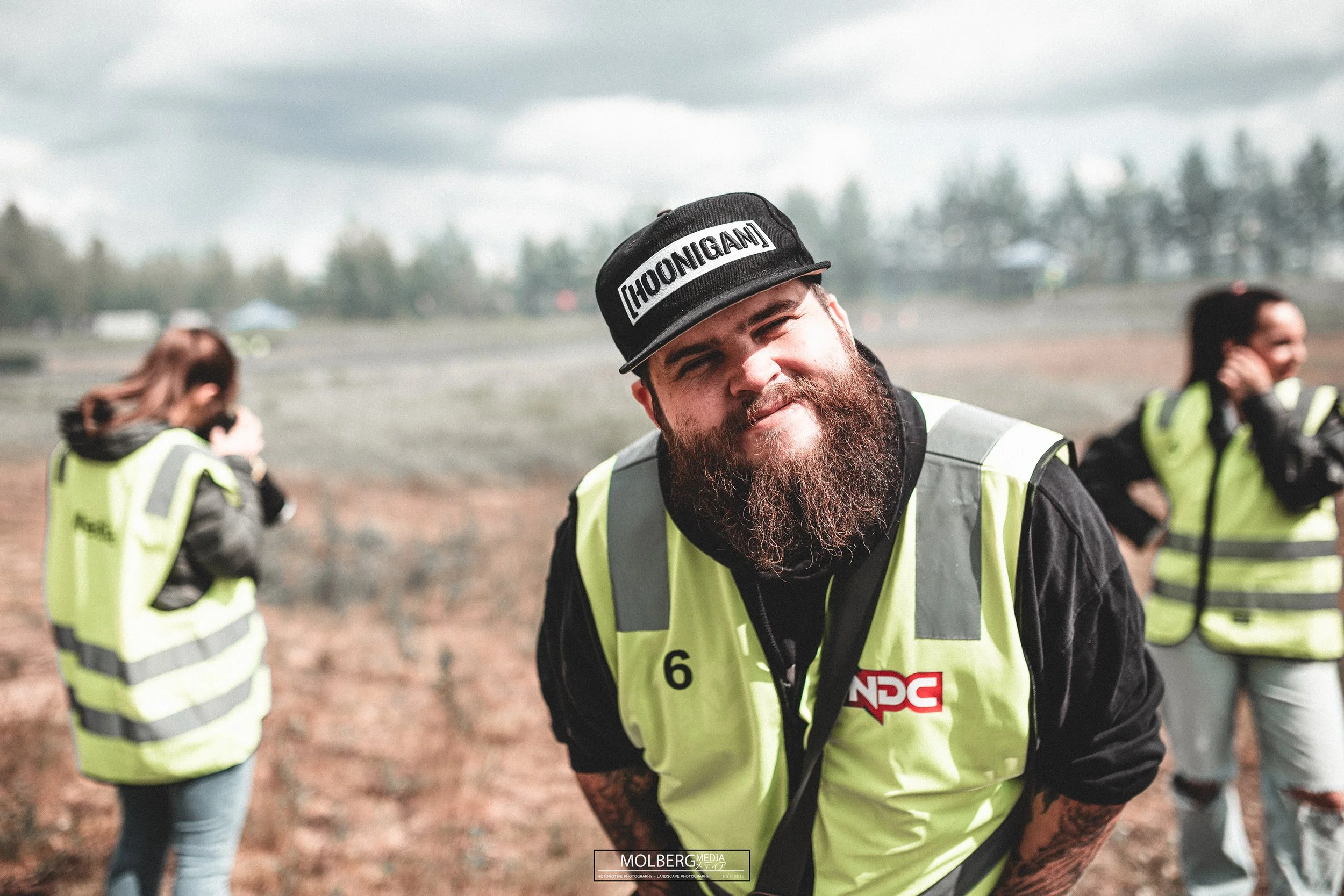 A bearded man wearing a black cap and a neon green safety vest, smiling at the camera, with two women in safety vests talking on their phones in the background outdoors on a cloudy day.