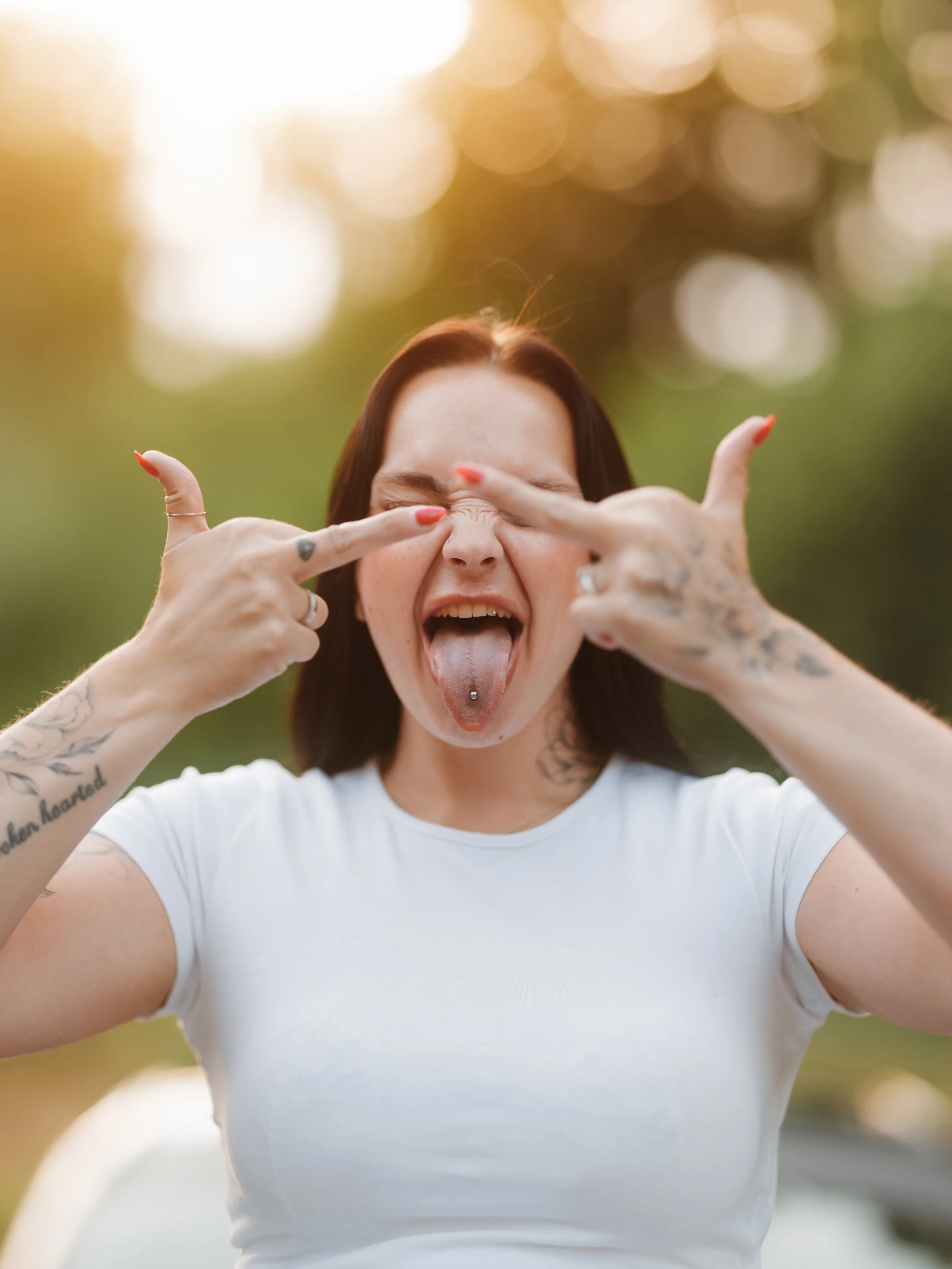 A woman with tattoos and a tongue piercing making a funny face outdoors, pulling her eyelids closed and sticking out her tongue, with a blurred background of green trees and sunlight.