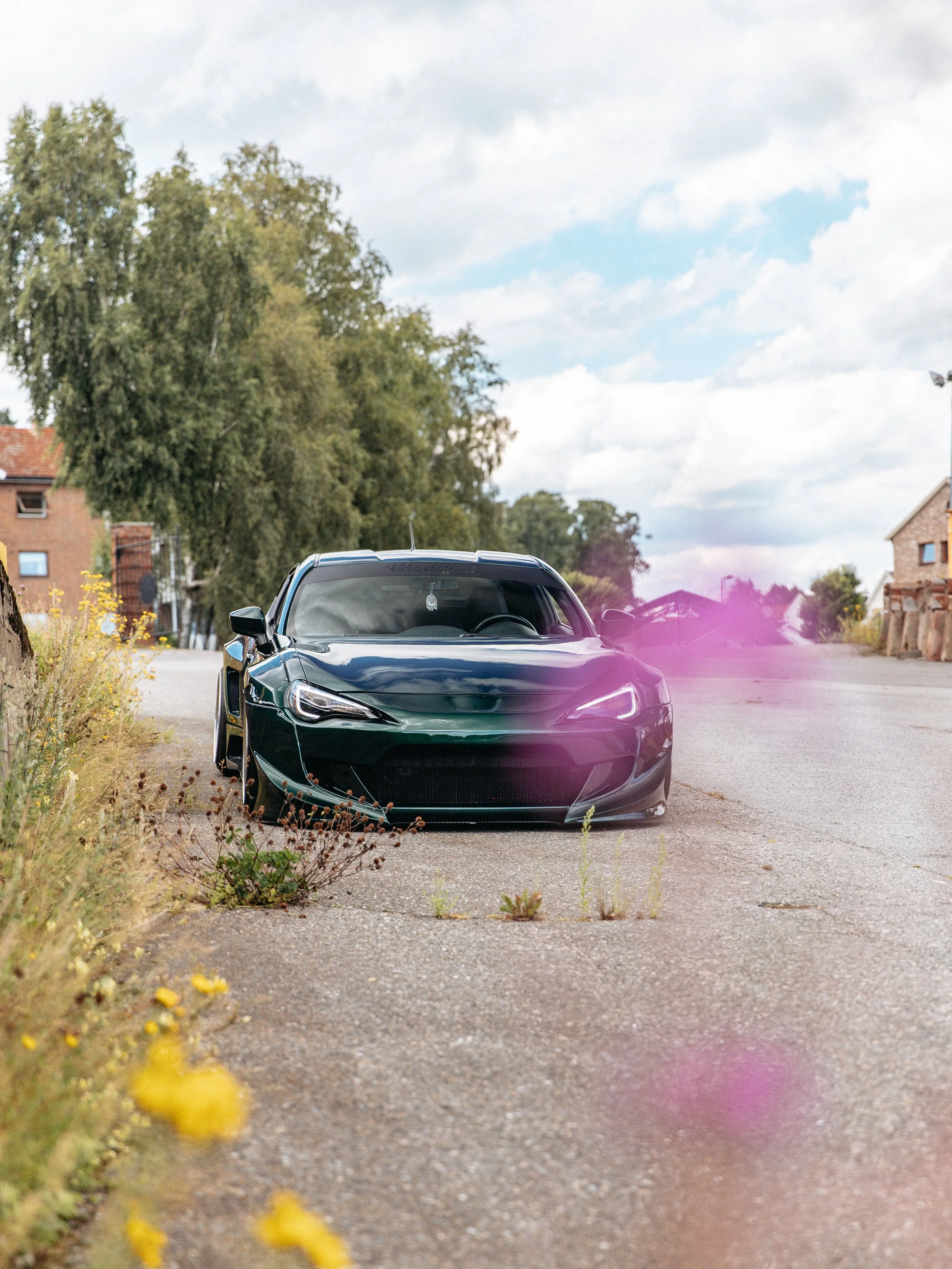 Black sports car parked on a roadside with trees and houses in the background, and a partly cloudy sky above.