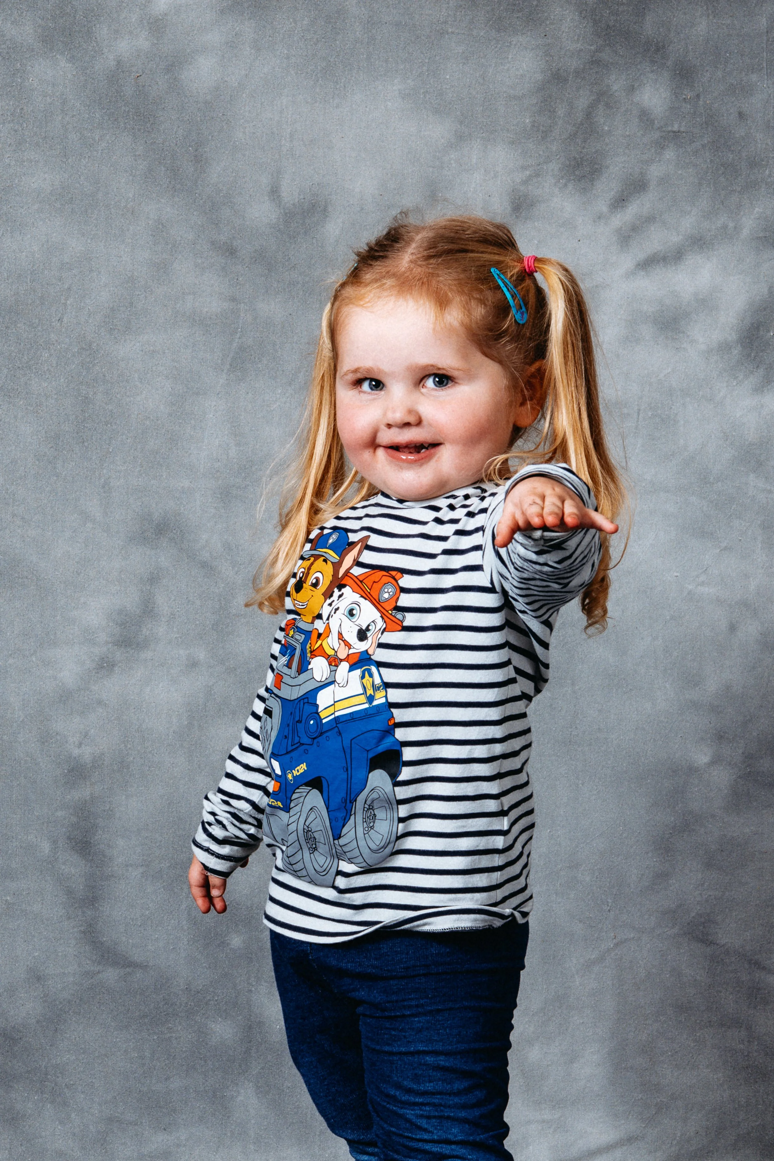 A young girl with red hair, blue eyes, and a big smile, wearing a striped shirt featuring characters from 'Paw Patrol', poses with one arm extended forward against a gray background.
