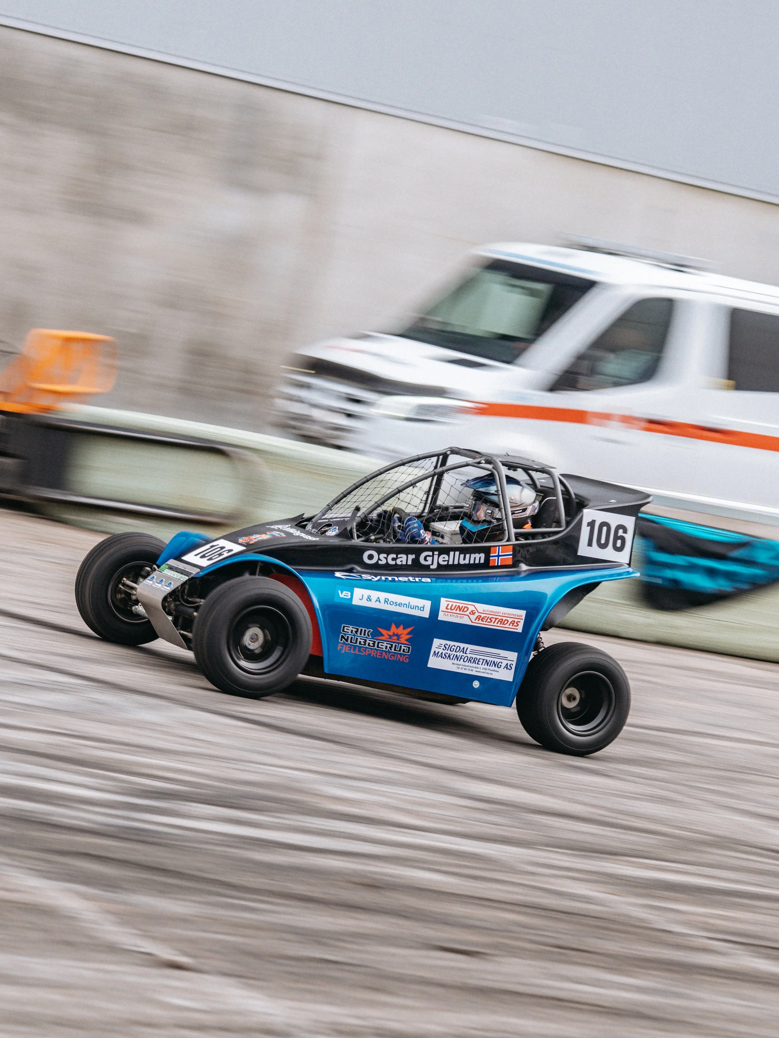 A blue and black off-road racing vehicle with the number 106 on it, driven by Oscar Gjellum, speeding on a dirt track with a safety barrier and a white emergency vehicle in the background.