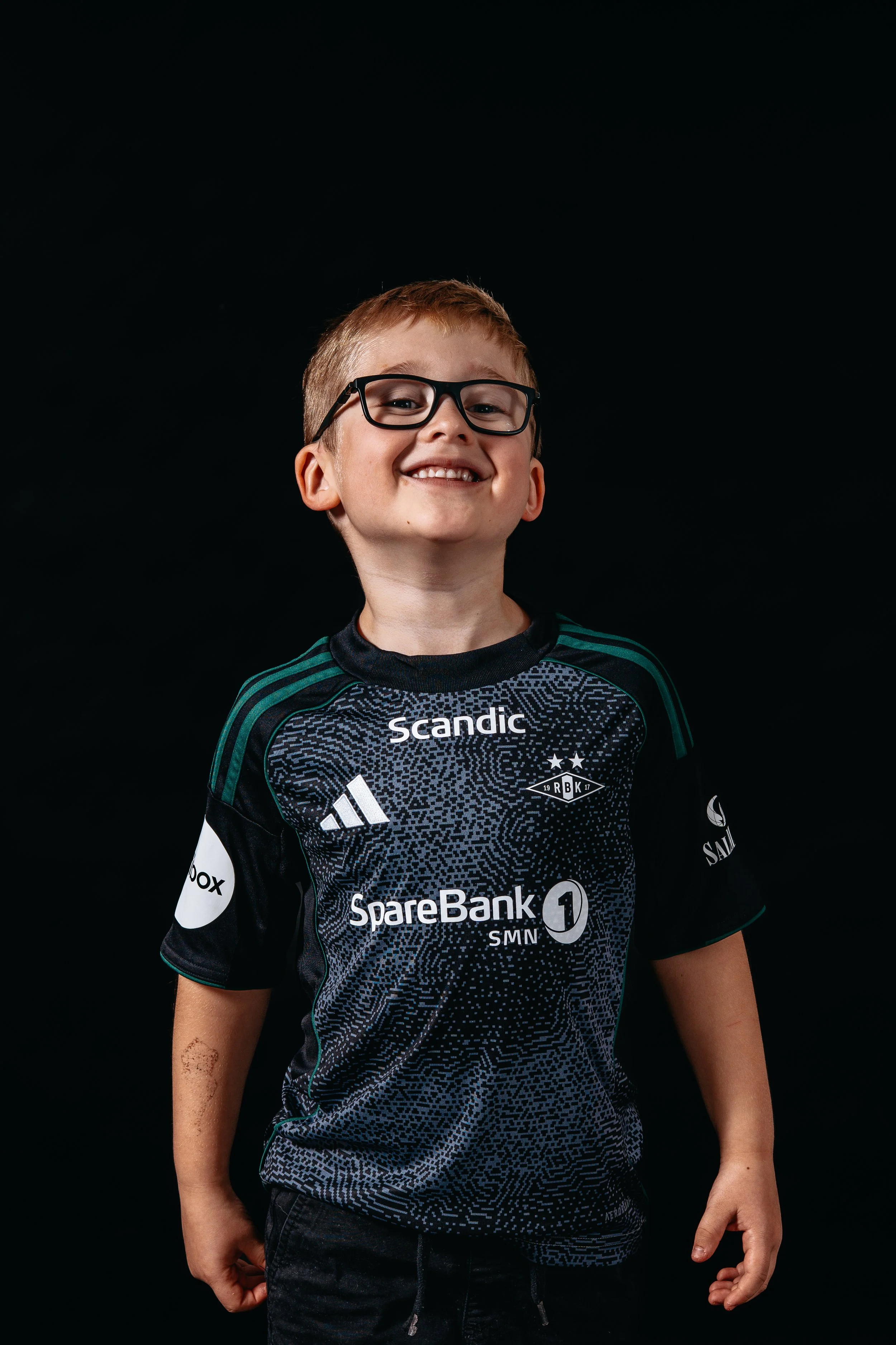 A smiling young boy with glasses, wearing a black sports jersey with sponsorship logos, standing against a black background.
