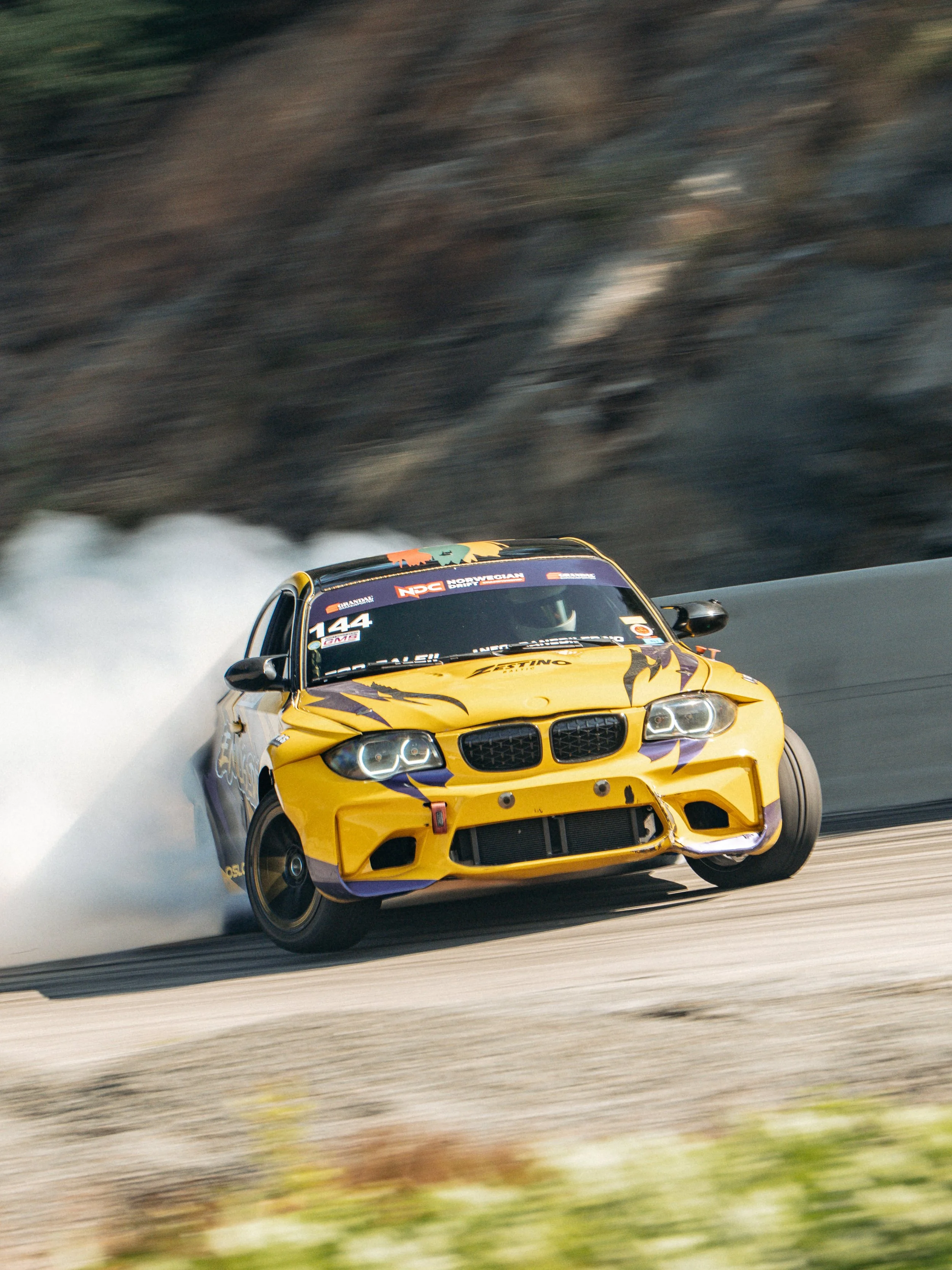 A yellow race car with purple accents, number 144, drifting on a race track with white smoke behind it and a rocky hillside in the background.