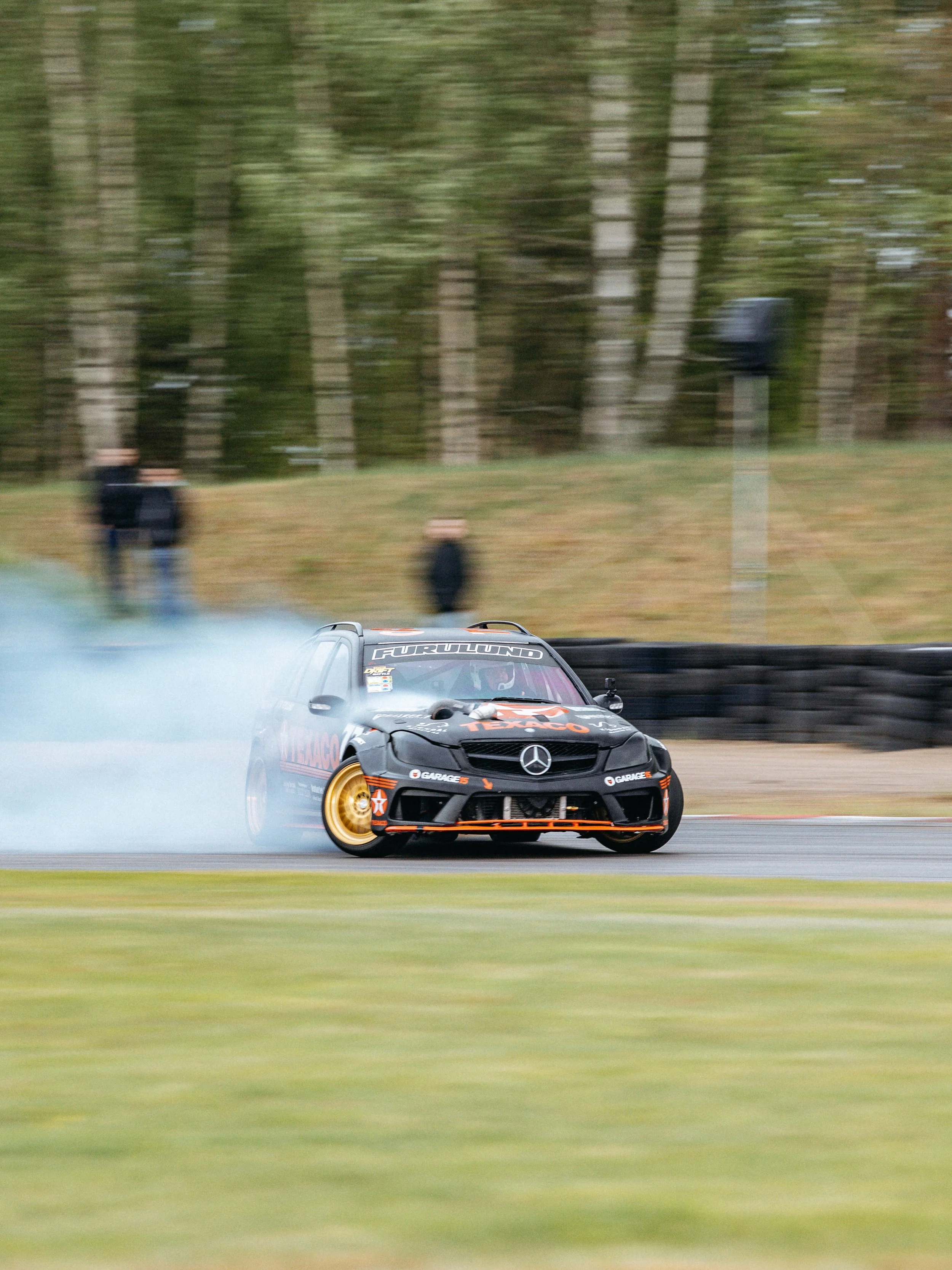 A black race car with gold wheels and orange accents drifting on a race track, smoke coming from the tires, with trees and blurred spectators in the background.