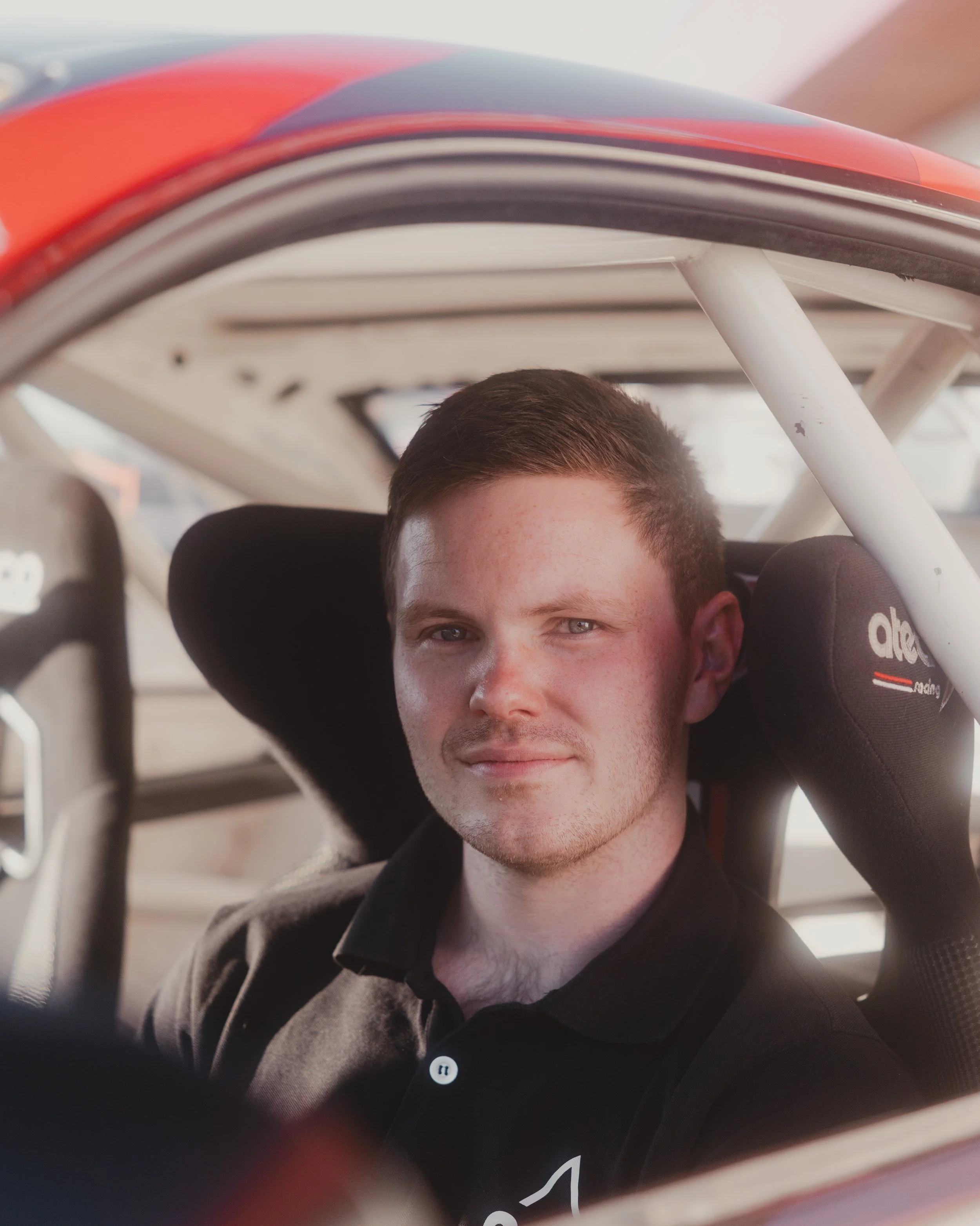 Close-up of a young man with short brown hair seated inside a race car, wearing a black polo shirt, with a confident expression.