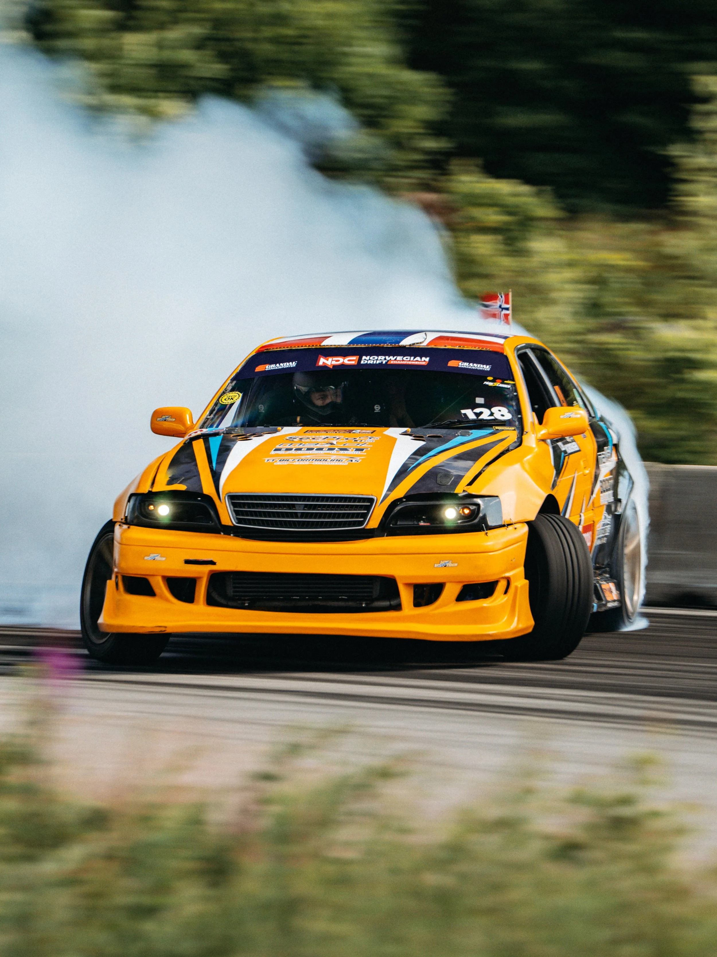 A yellow race car speeding on a track, creating smoke behind it, with a driver visible inside, and a small Norwegian flag on the rear wing.