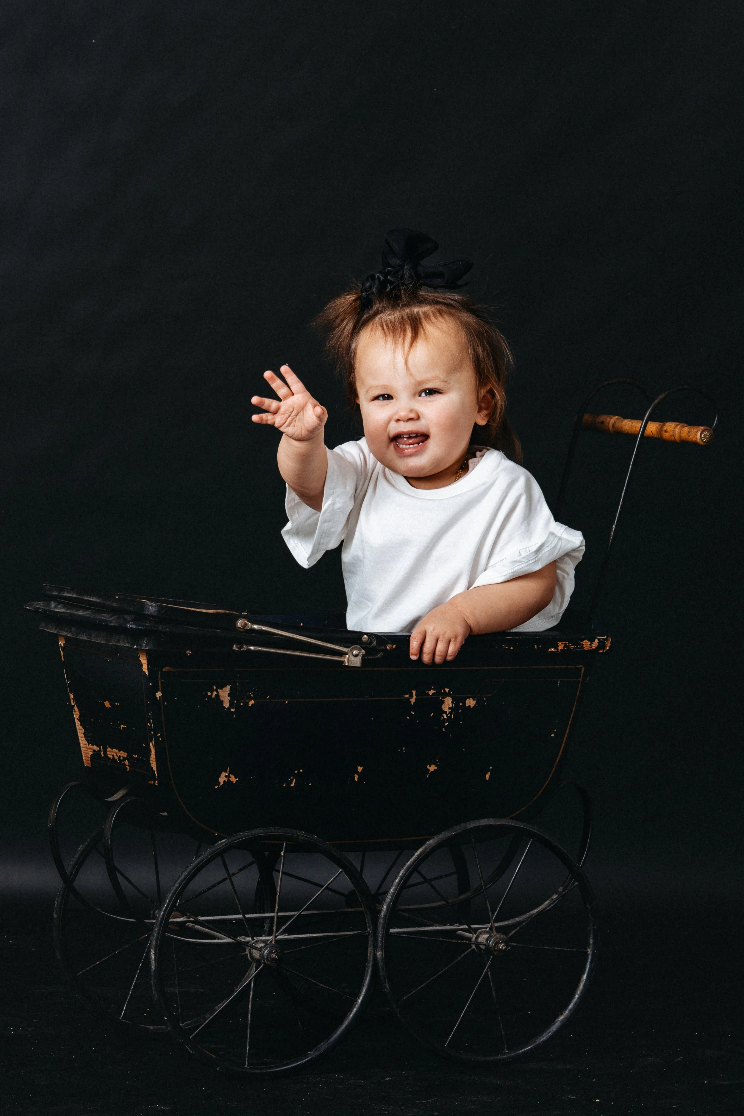 A young child with a black bow in her hair sitting in an antique wooden doll stroller against a black background, waving and smiling.