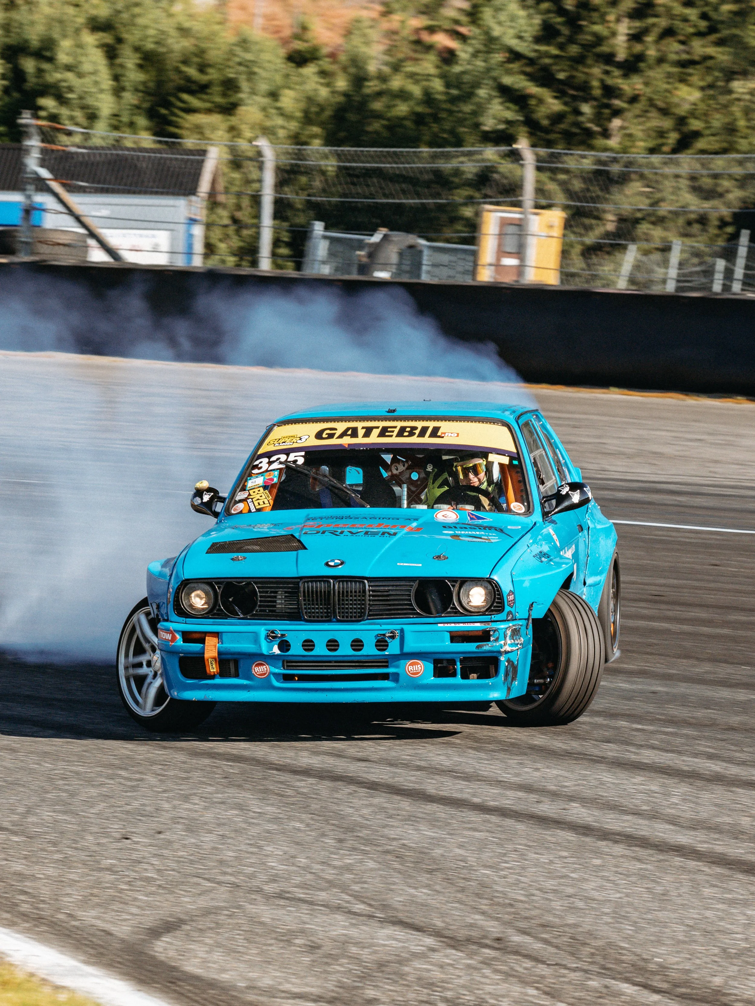 A blue BMW race car with visible damage on the front bumper speeding on a racetrack, with smoke coming from the rear tires, and trees in the background.