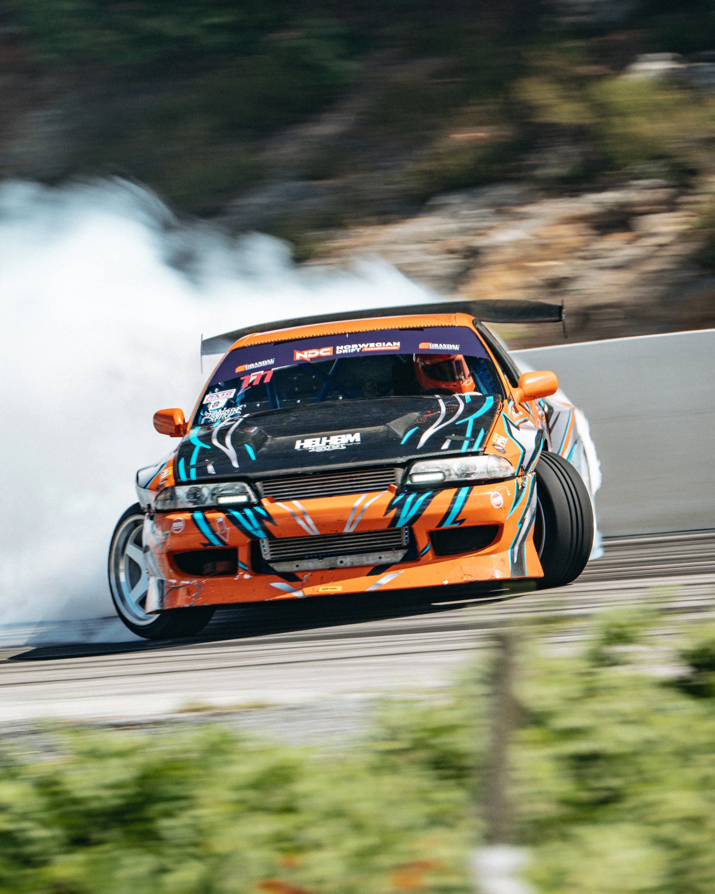 A race car with orange, black, and blue graphics drifting on a race track, producing tire smoke.
