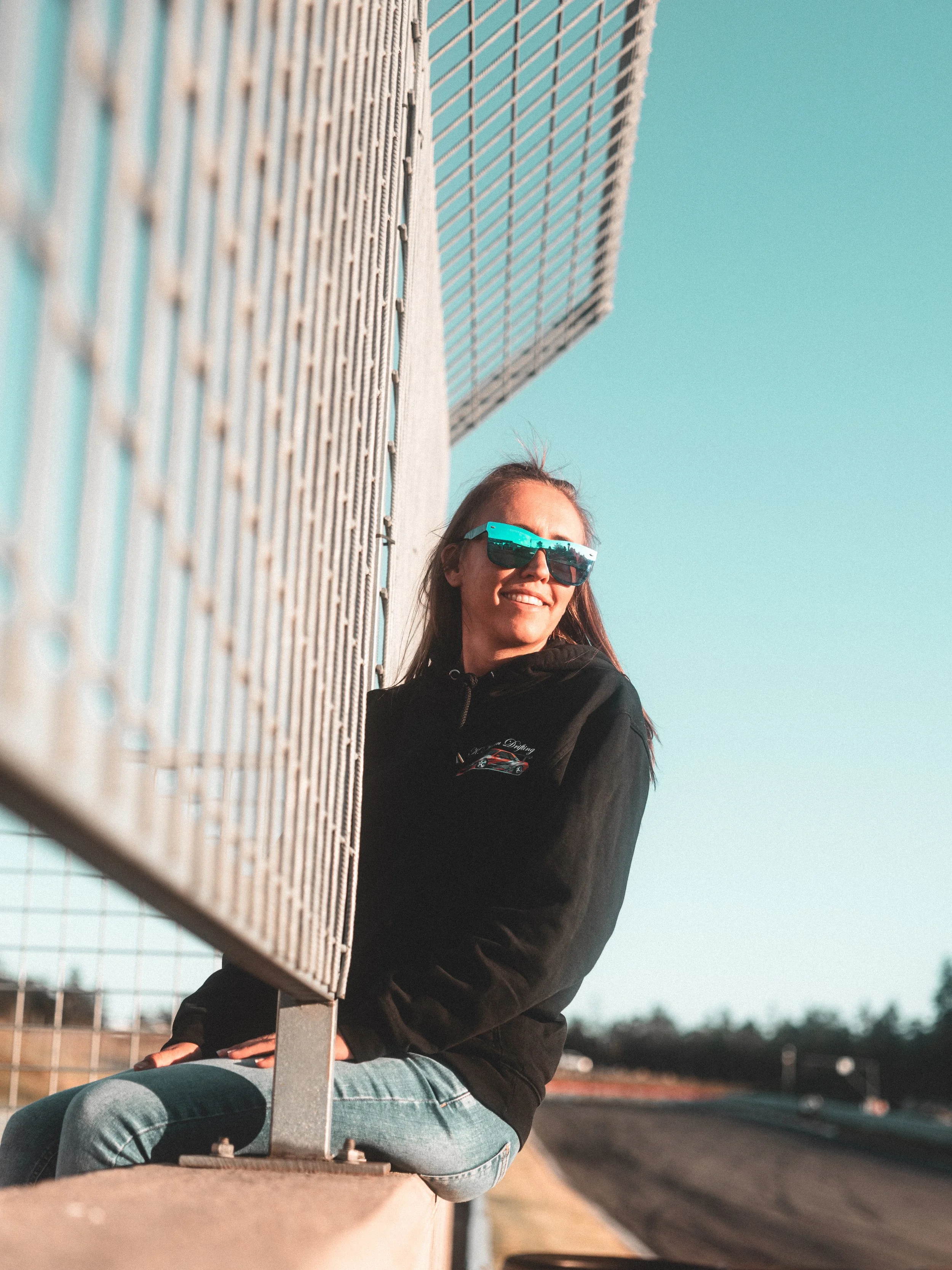 A young woman sitting on a barrier wall at a race track, wearing sunglasses and a black hoodie, smiling as she looks down the track during daytime.