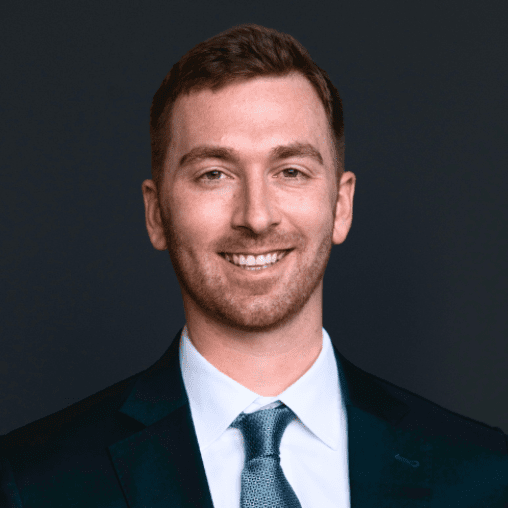 Professional headshot of a young man in a suit and tie, smiling against a dark background.