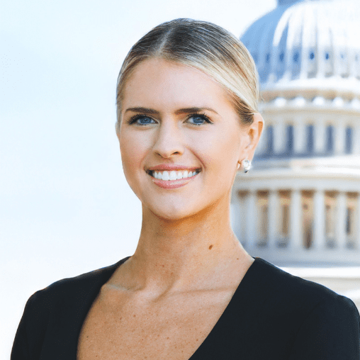 A woman with blonde hair smiling in front of a government building with a dome.