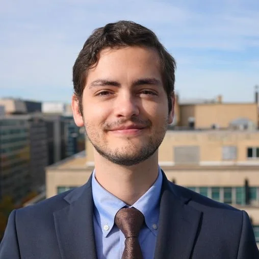 A young man with short dark hair and a light beard, dressed in a dark suit jacket, light blue shirt, and brown tie, smiling outdoors with a cityscape background under a partly cloudy sky.