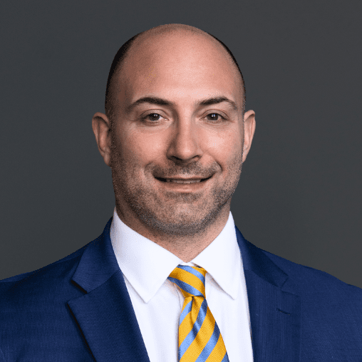 Man wearing a blue suit, white shirt, and yellow striped tie, smiling against a gray background.