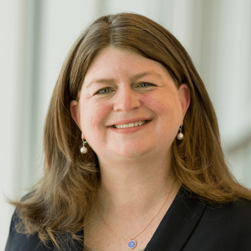 A smiling woman with shoulder-length brown hair, wearing pearl earrings and a black blazer, standing in front of a light-colored background.