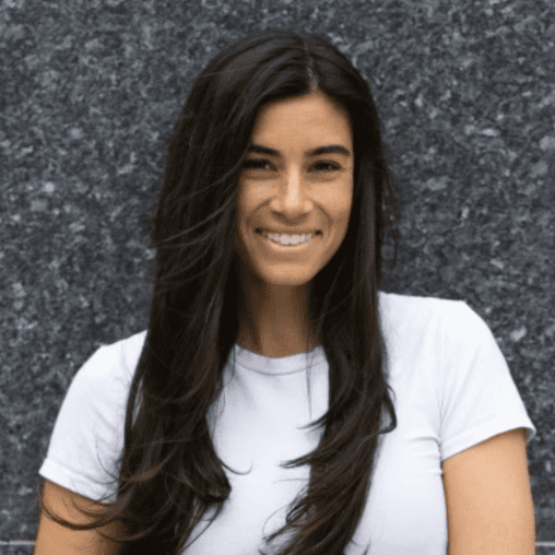 A woman with long dark hair wearing a white t-shirt standing in front of a dark textured background.