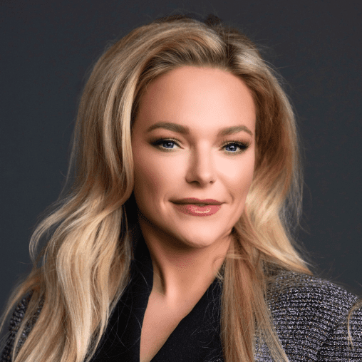 Portrait of a woman with long blonde hair, wearing a dark blazer, smiling slightly against a dark background.