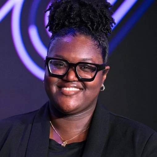 A woman with glasses and braided hair styled in a tall bun, smiling, wearing a black blazer and jewelry, against a dark background with purple neon lights.