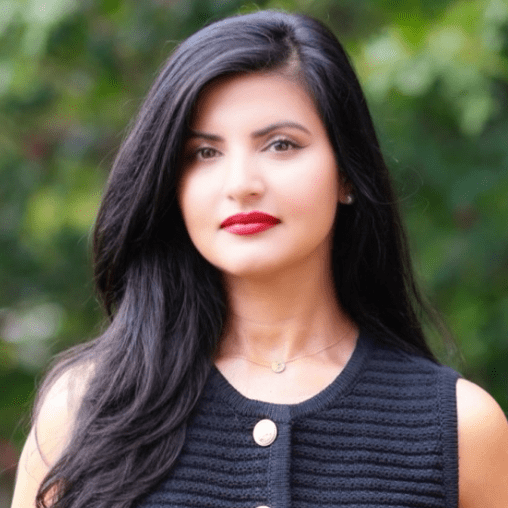 A woman with long, dark hair wearing a sleeveless navy blue top outdoors with blurred green foliage in the background.