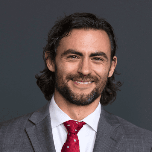 A portrait of a smiling man with dark hair and a beard, wearing a gray suit, white shirt, and red tie, against a dark gray background.