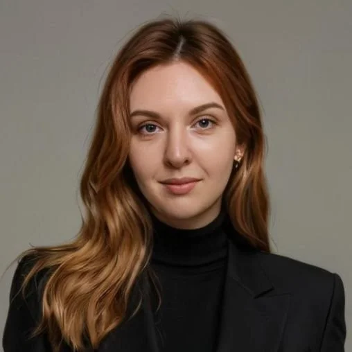Portrait of a young woman with long wavy red hair wearing a black blazer and turtleneck, neutral background.