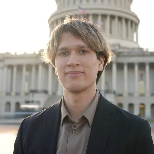 A young man with light brown hair and a slight smile standing outdoors in front of a government building, possibly the U.S. Capitol, during the daytime.