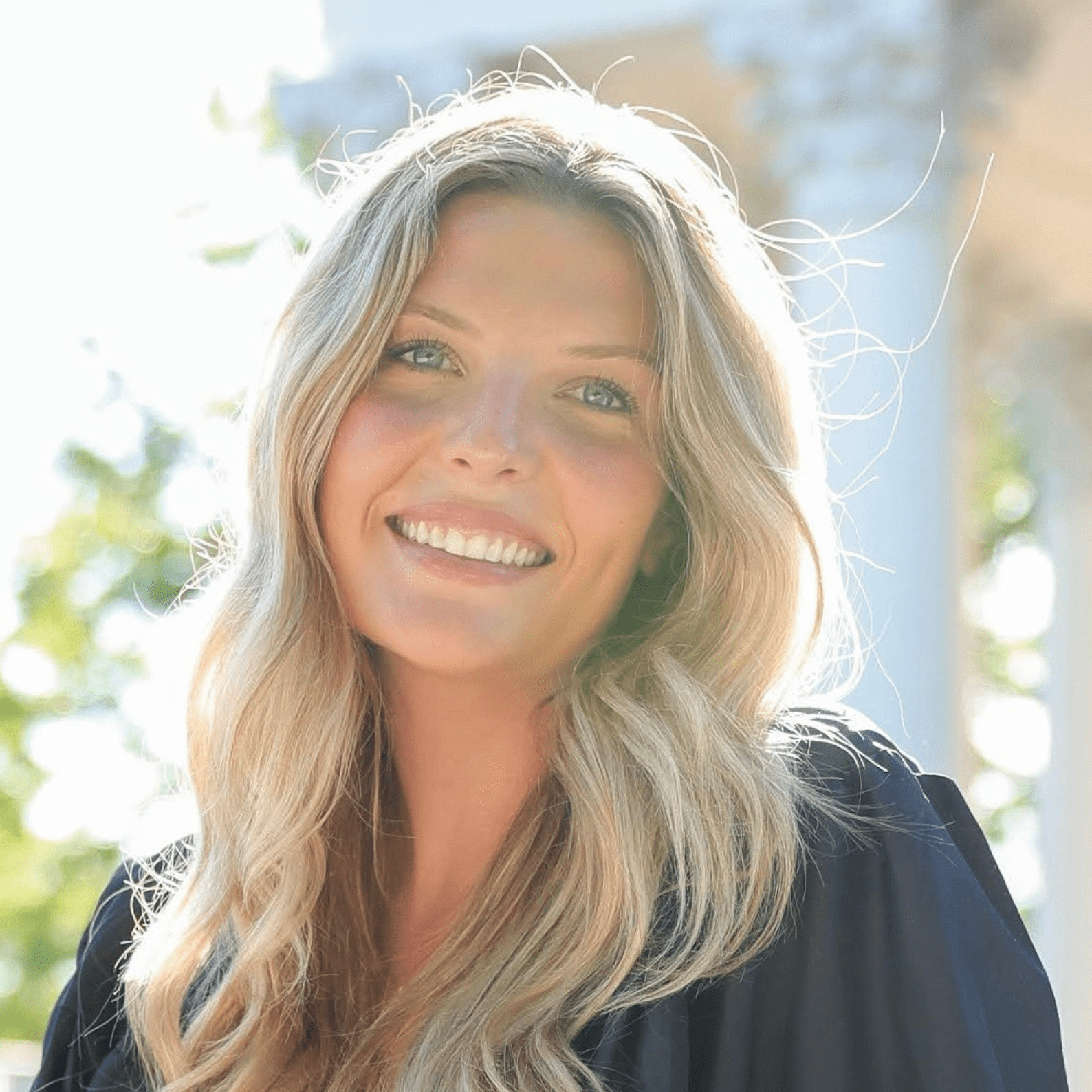 A smiling woman with long blonde hair, blue eyes, and light skin, outdoors with sunlight and blurred greenery in the background.