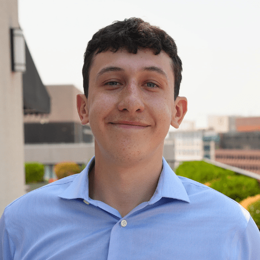 Young man with short dark hair wearing a light blue button-up shirt, smiling outdoors with blurred buildings and greenery in the background.