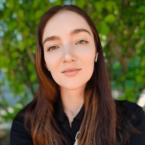 A young woman with long brown hair wearing a black shirt, standing outdoors with green foliage in the background.