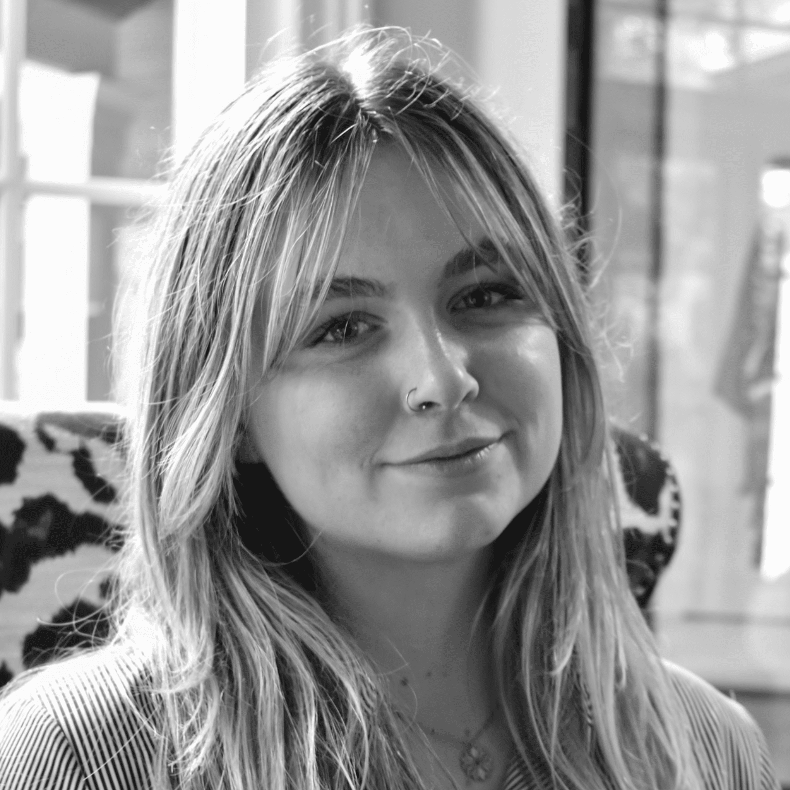 Black and white photo of a young woman with long hair, smiling softly, wearing a nose ring and a necklace, seated indoors near a window.