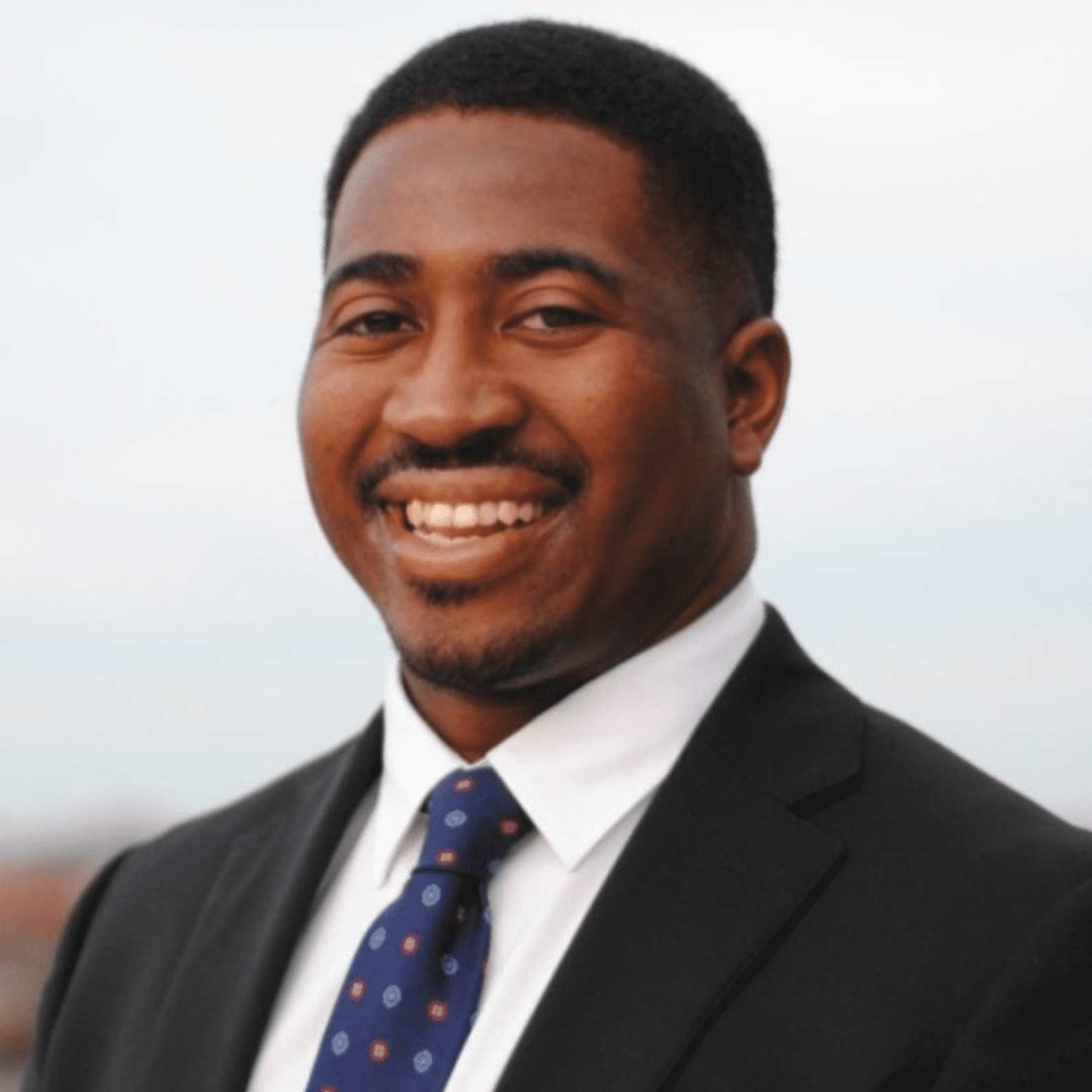 A young Black man in a business suit smiling outdoors with a blurred sky background.