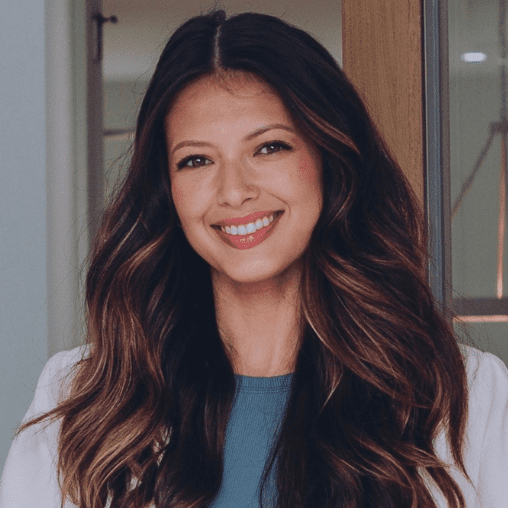 A woman with long, wavy brown hair smiling at the camera in an indoor setting.