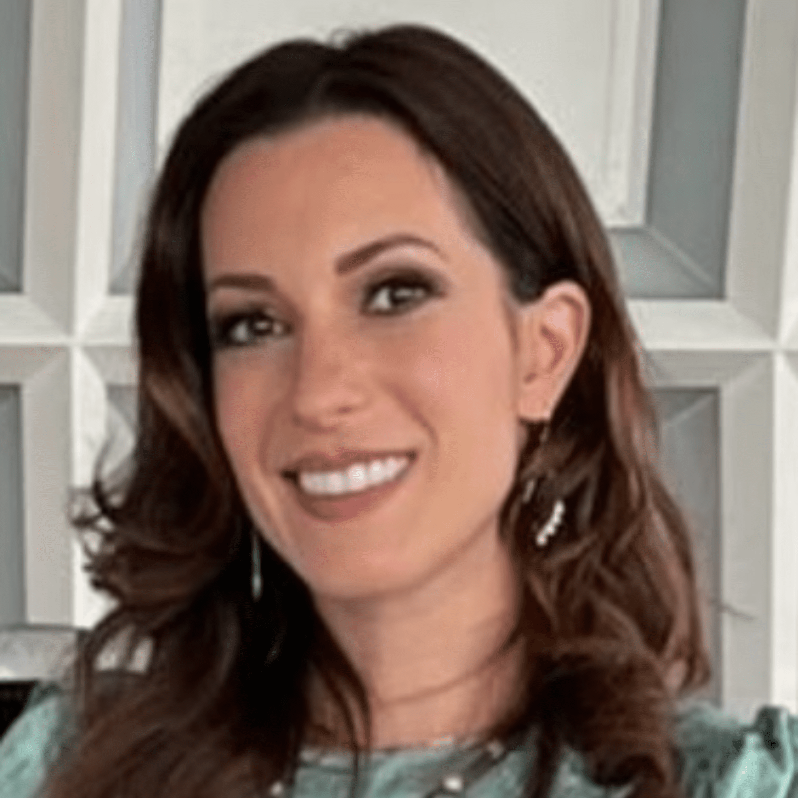 A woman with wavy brown hair smiling in front of a geometric-patterned wall.
