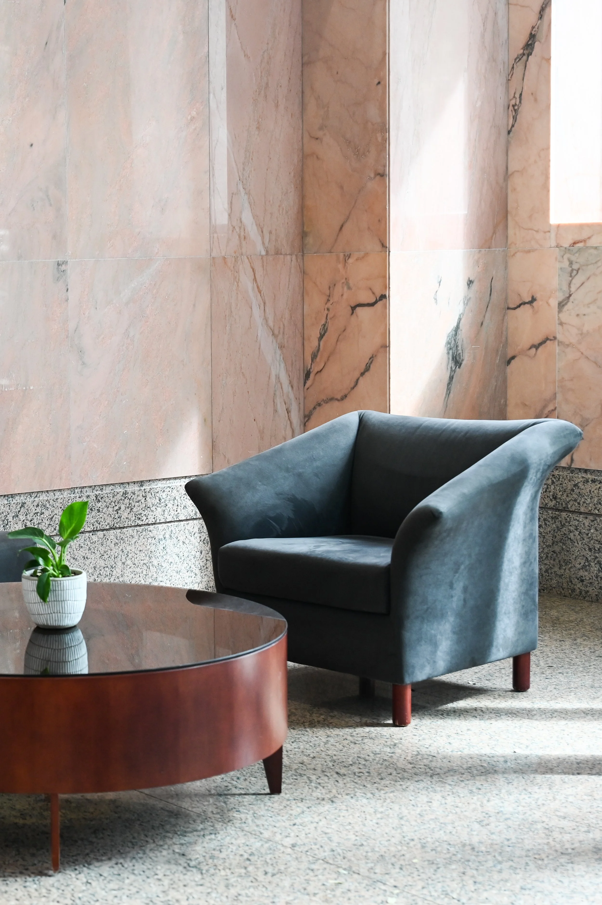 A blue velvet armchair and a round wooden coffee table with a small white potted plant, set against a pink marble wall in a modern interior.