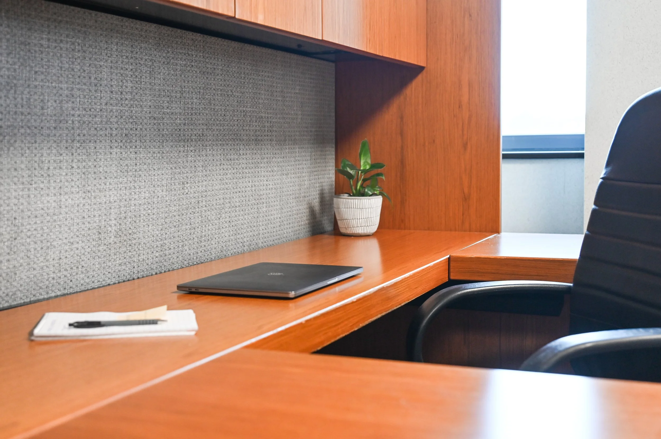A wooden office desk with a closed laptop, a notebook with a pen, a small potted plant, and a black office chair.