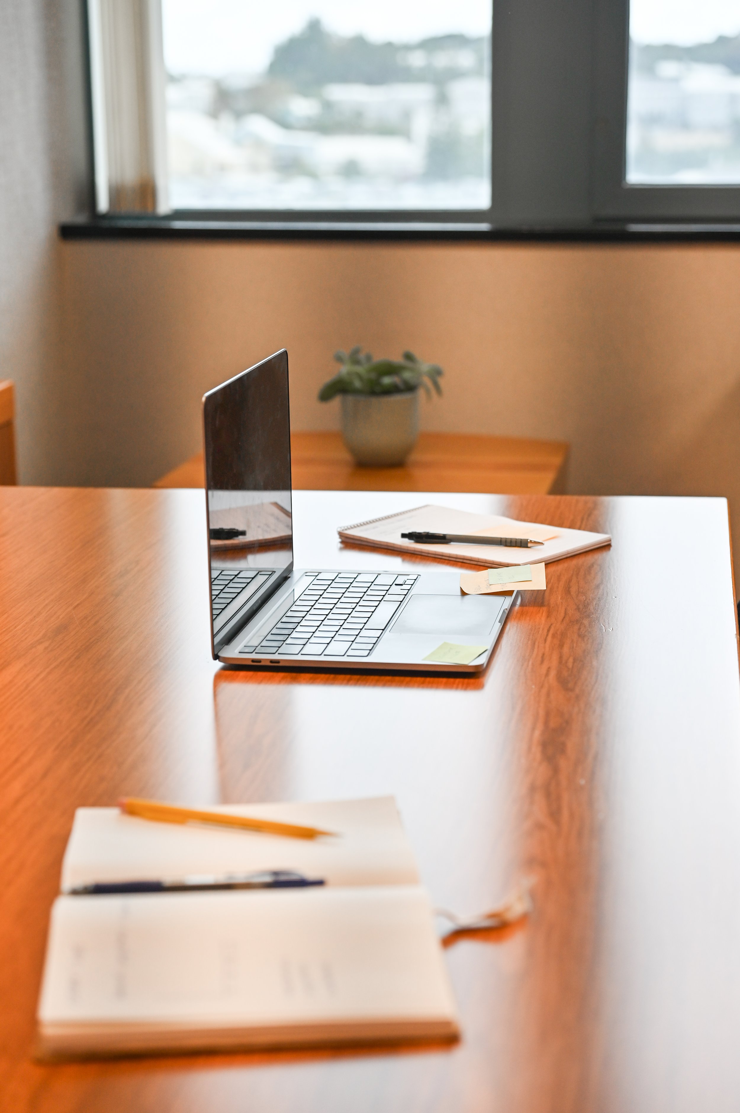 A wooden desk with an open laptop, notebooks, pens, sticky notes, and a blurred potted plant near a window in a well-lit office.