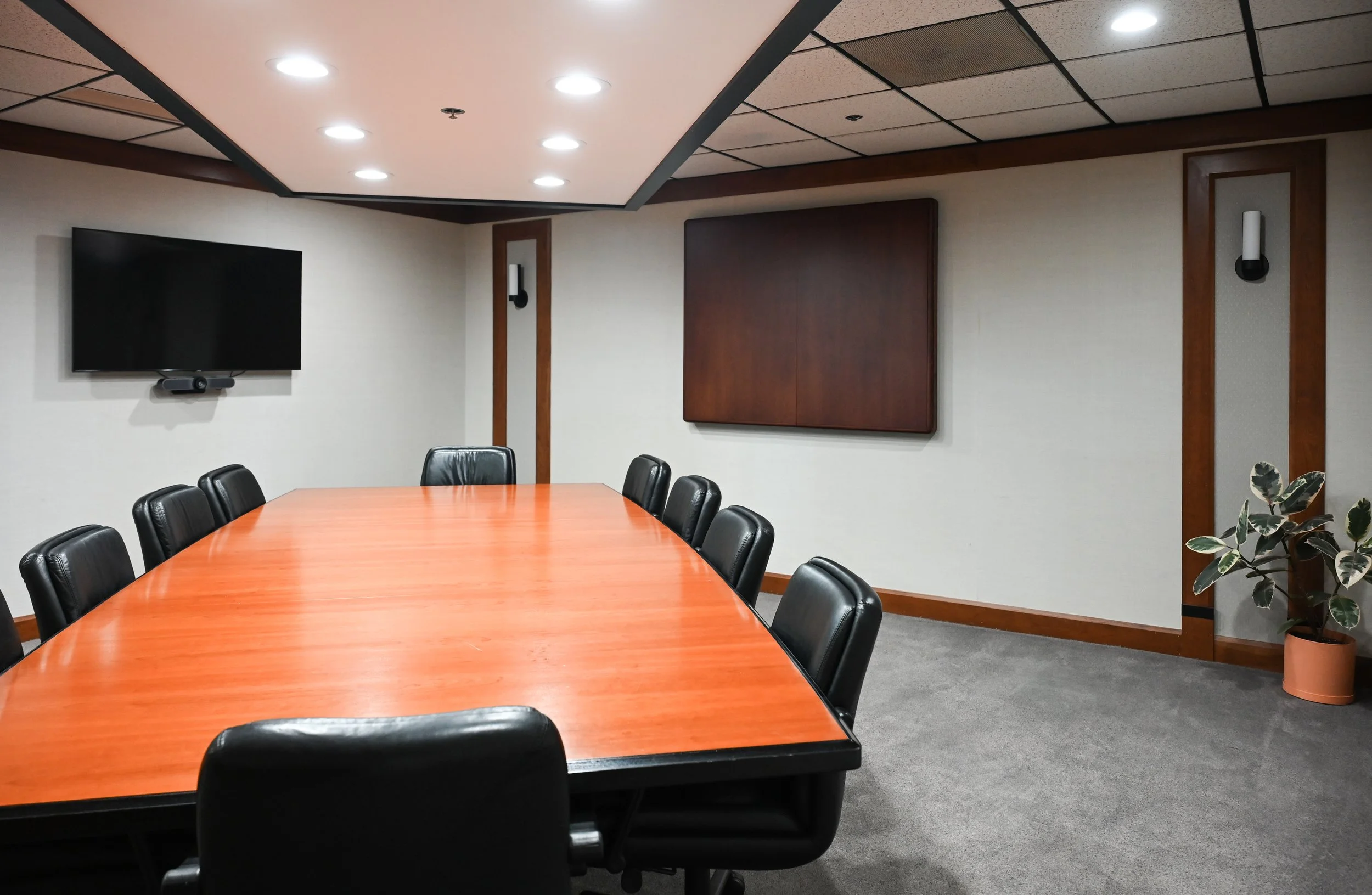 A conference room with a large wooden table surrounded by black leather chairs, a wall-mounted flat-screen TV, a wall-mounted display or panel, and a potted plant in the corner.