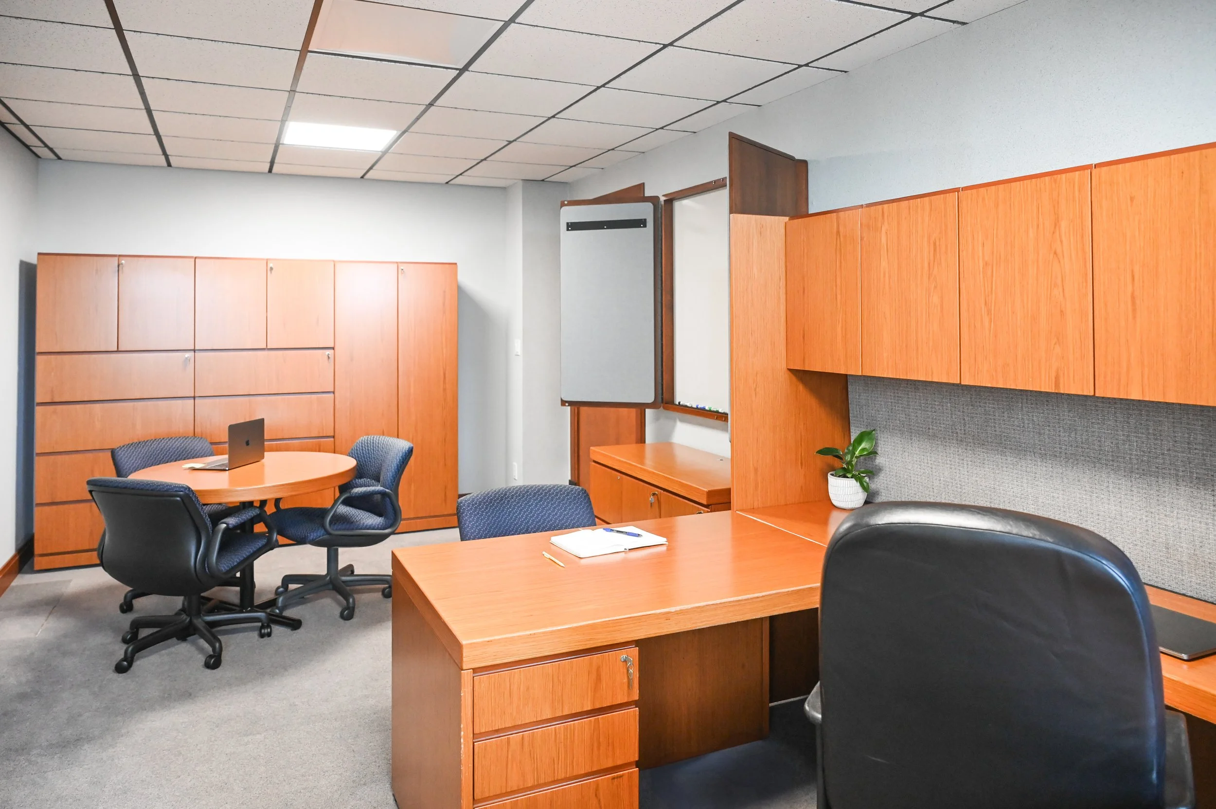 An office room with wooden desks, black office chairs, a cabinet with drawers, a whiteboard, a gray whiteboard with a sliding cover, a small potted plant, and a round table with two chairs; ceiling tiles and carpeted floor.
