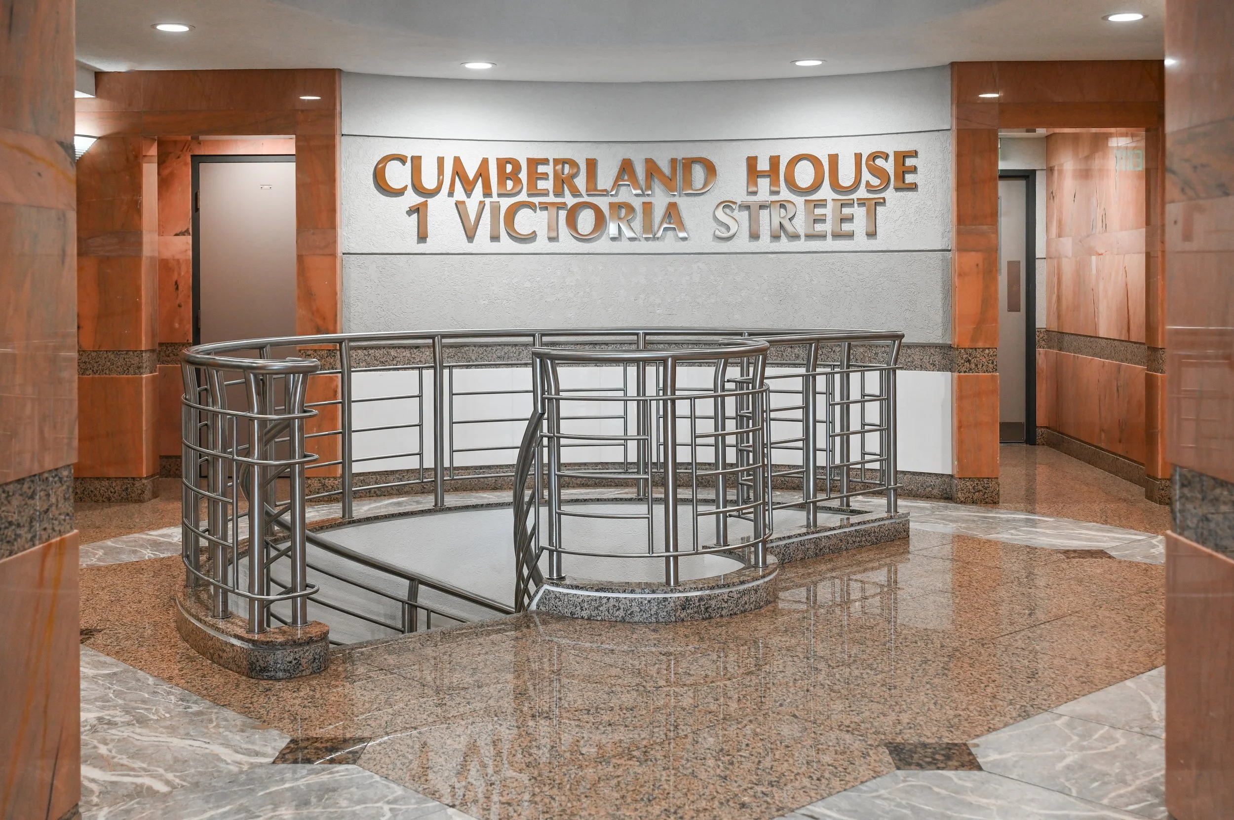 Indoor staircase leading down to a basement or lower level at Cumberland House, 1 Victoria Street, with metal railing, marble flooring, curved walls, and a sign on the wall.
