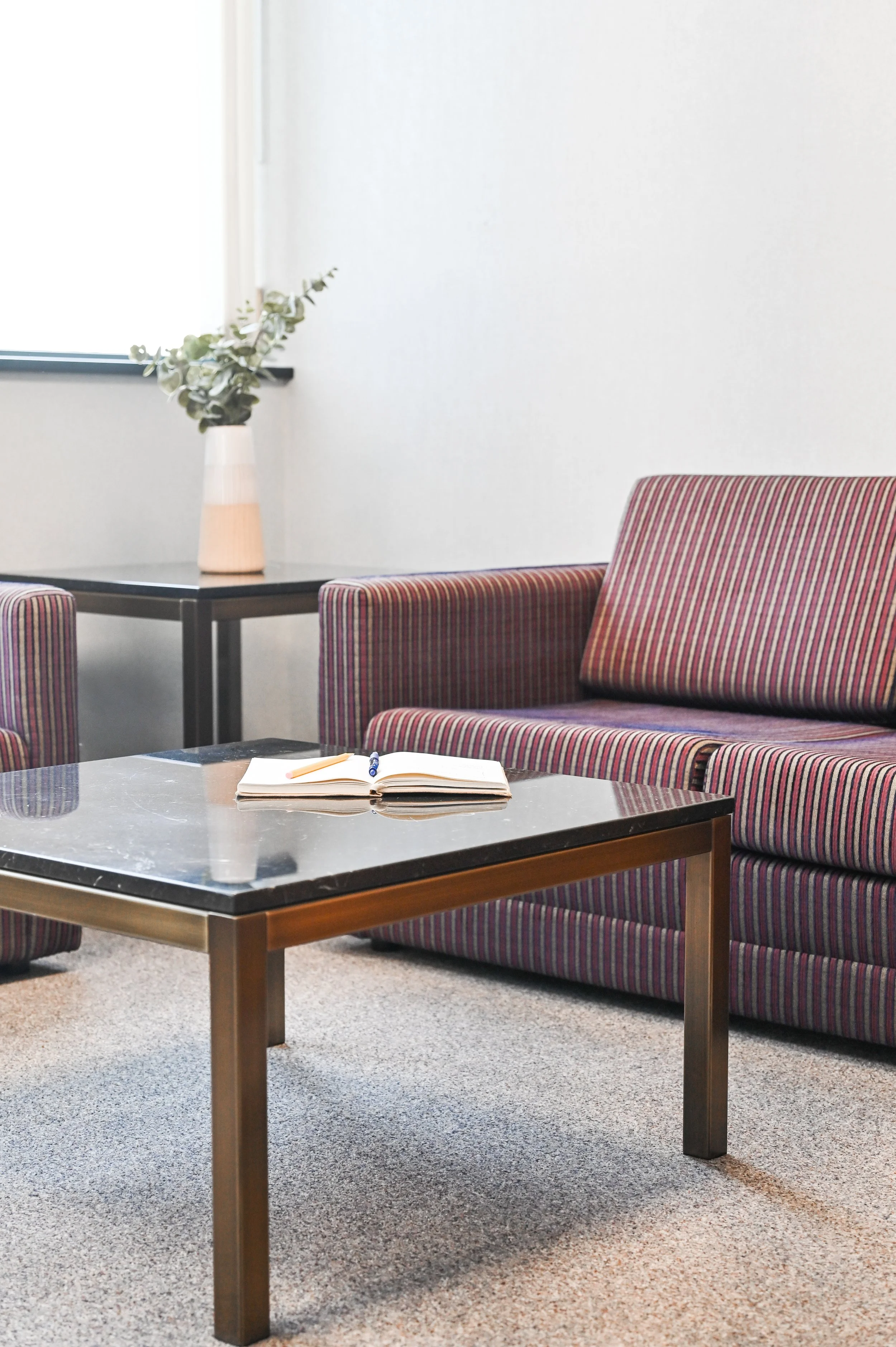 Modern office or lounge with a striped sofa, a glass-top coffee table with a notebook and pen, a side table with a vase of greenery, and a window with white walls.