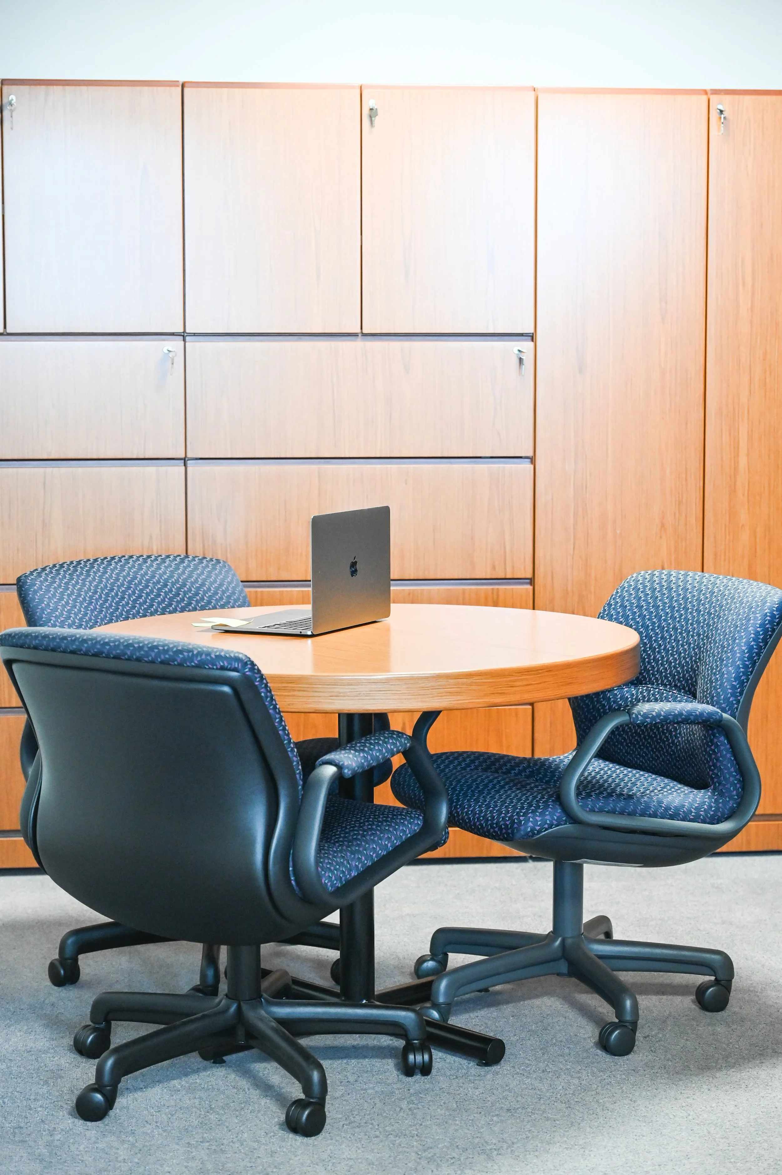 An office space with a round wooden table, two blue patterned chairs, and a closed laptop on the table, against a wooden cabinet background.