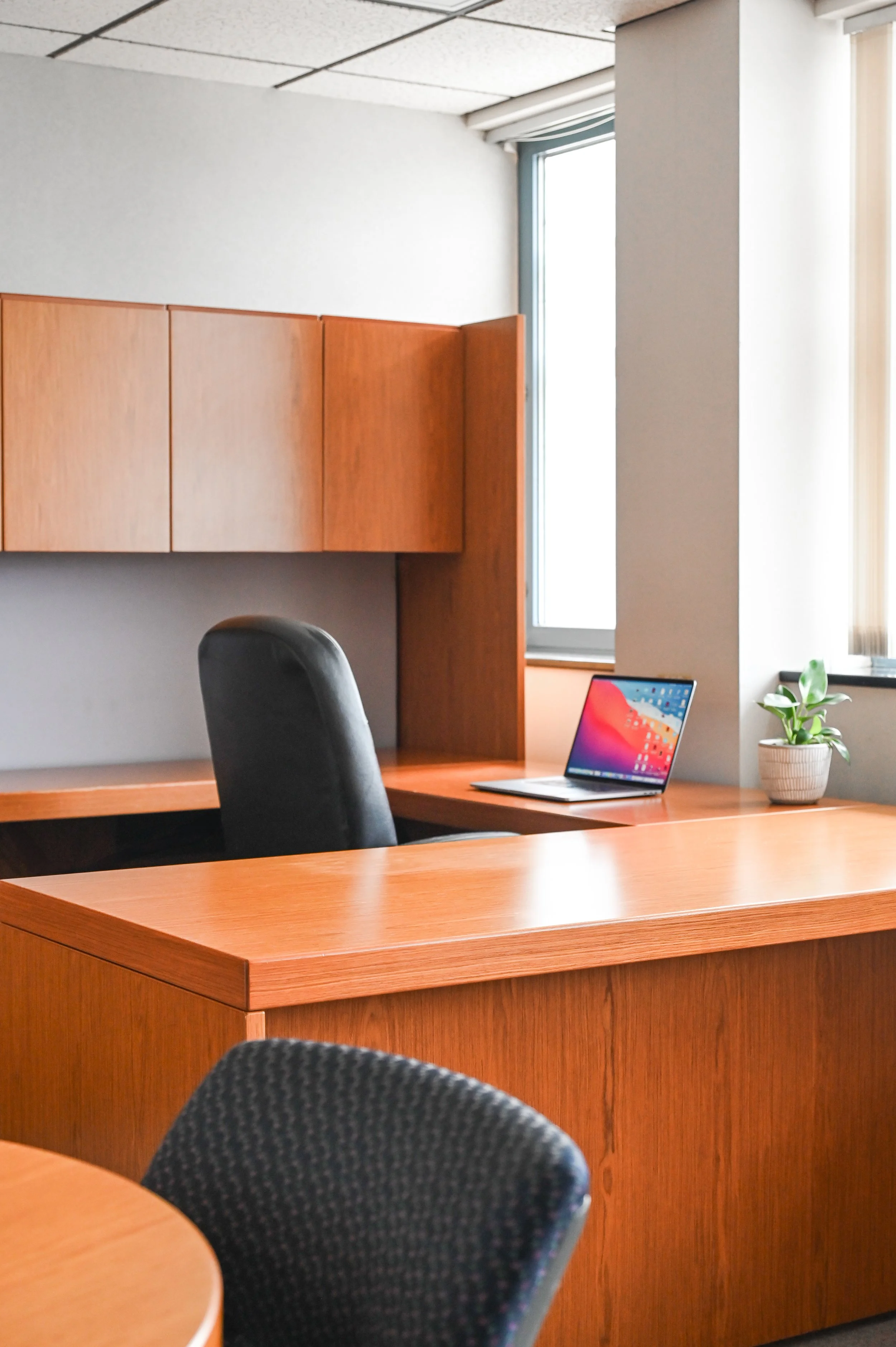An office with a wooden desk, a black chair, a laptop, a small plant, and wooden wall cabinets. There are windows with blinds letting in natural light.