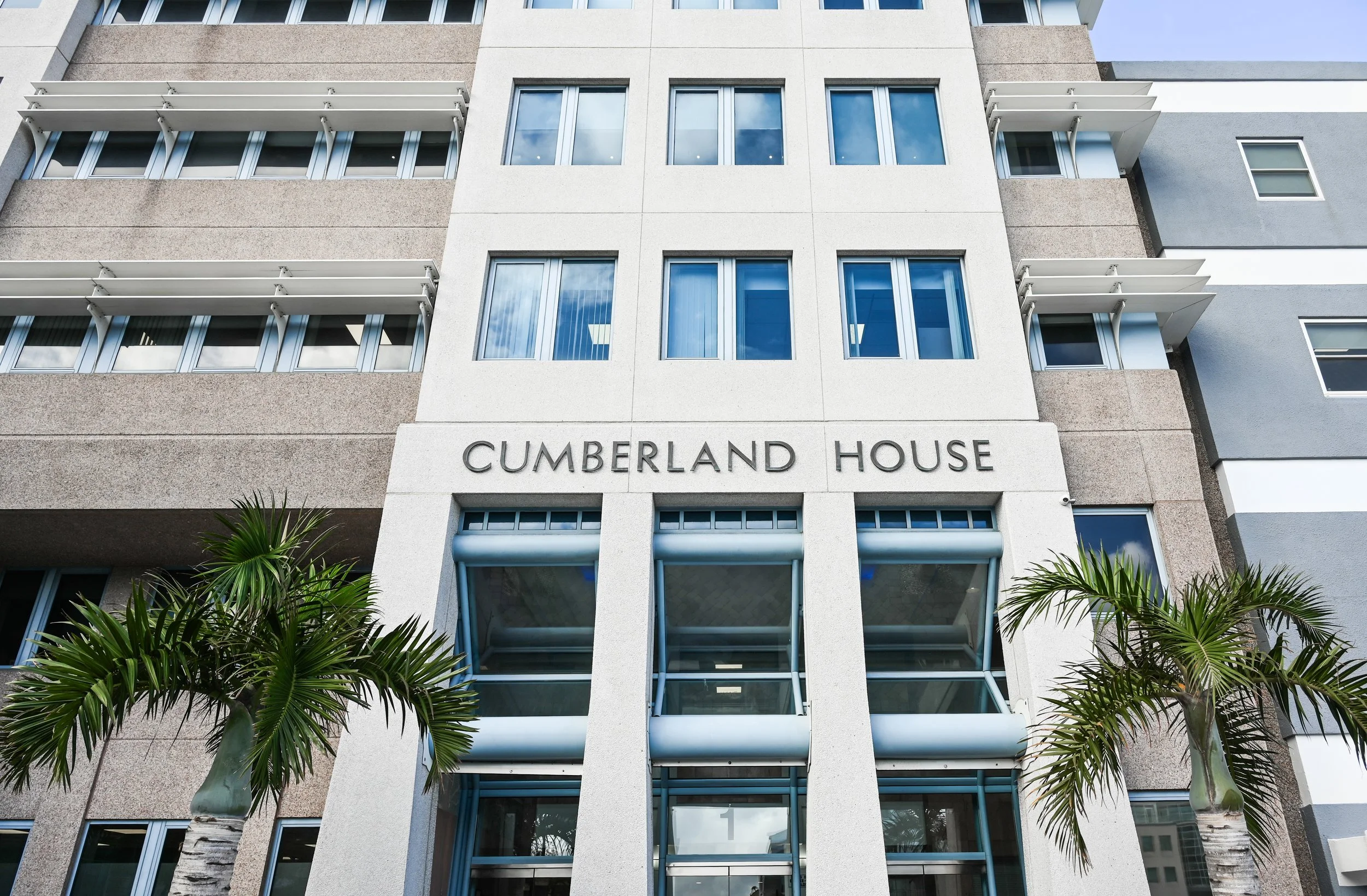 Front view of Cumberland House, a modern multi-story building with palm trees in front, large windows, and the building's name on the facade.