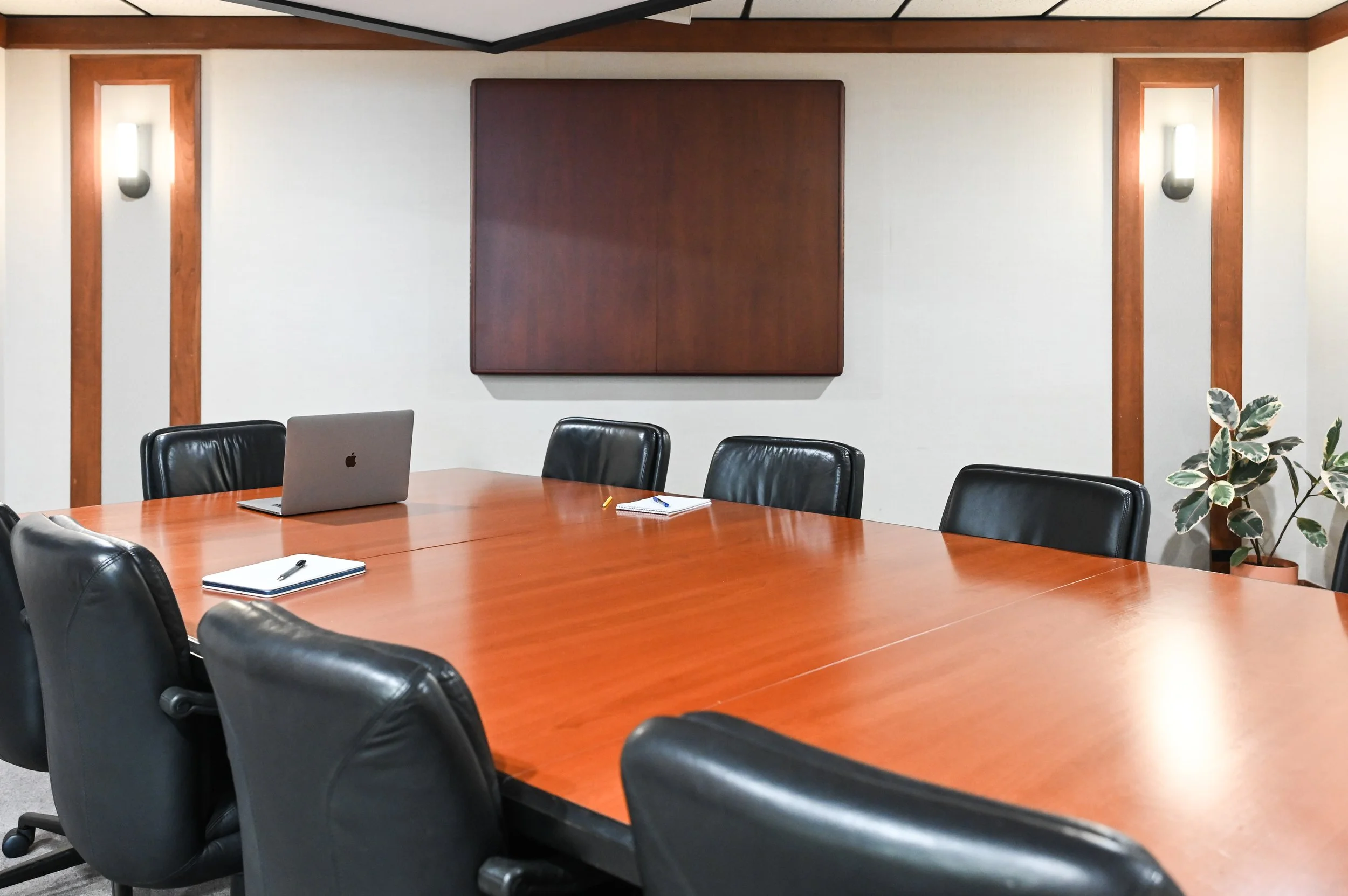 A conference room with a large wooden table, black leather chairs, a laptop, notebooks, and pens. The back wall has a large wooden panel and decorative wall lights. There is a potted plant in the corner.