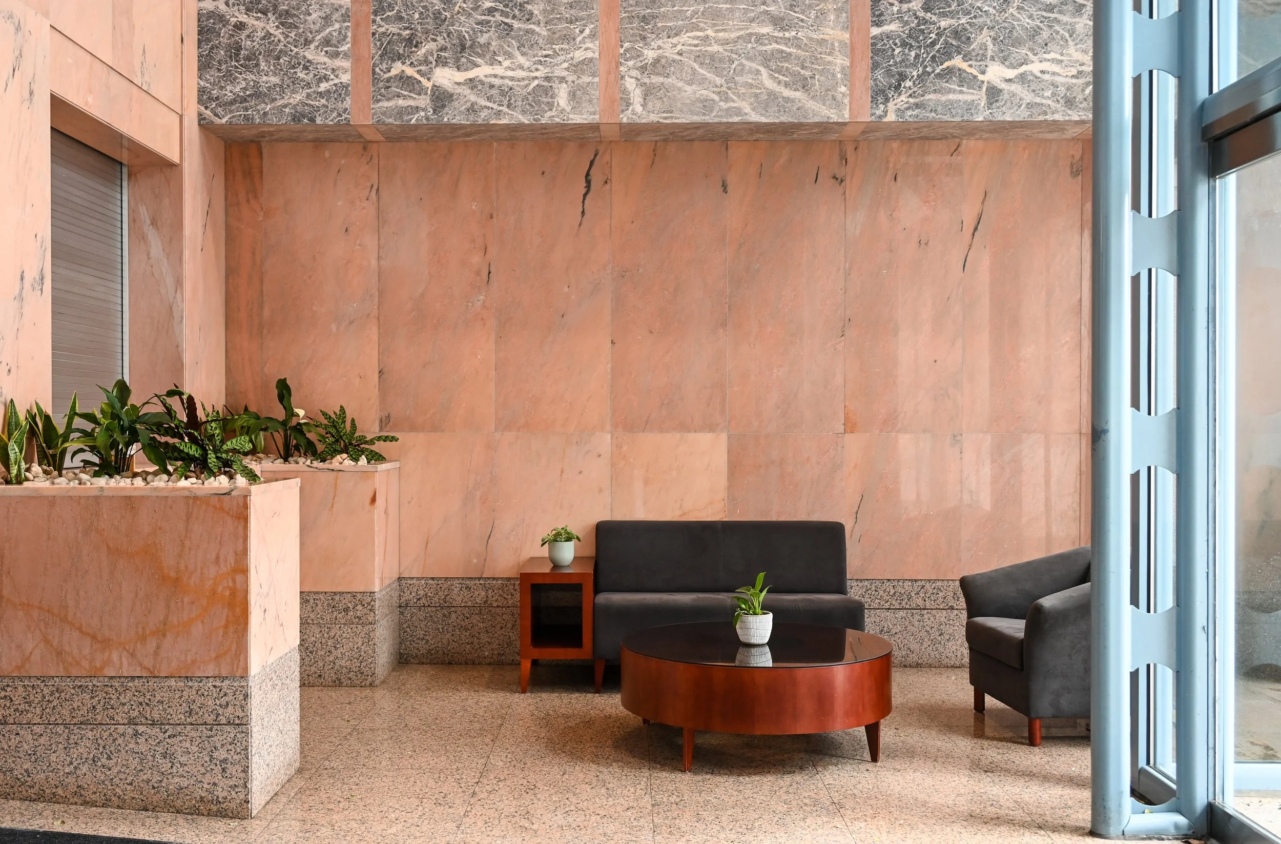 Lobby with pink marble walls, two gray chairs, a small wooden table with potted plants, and a blue Entry door.