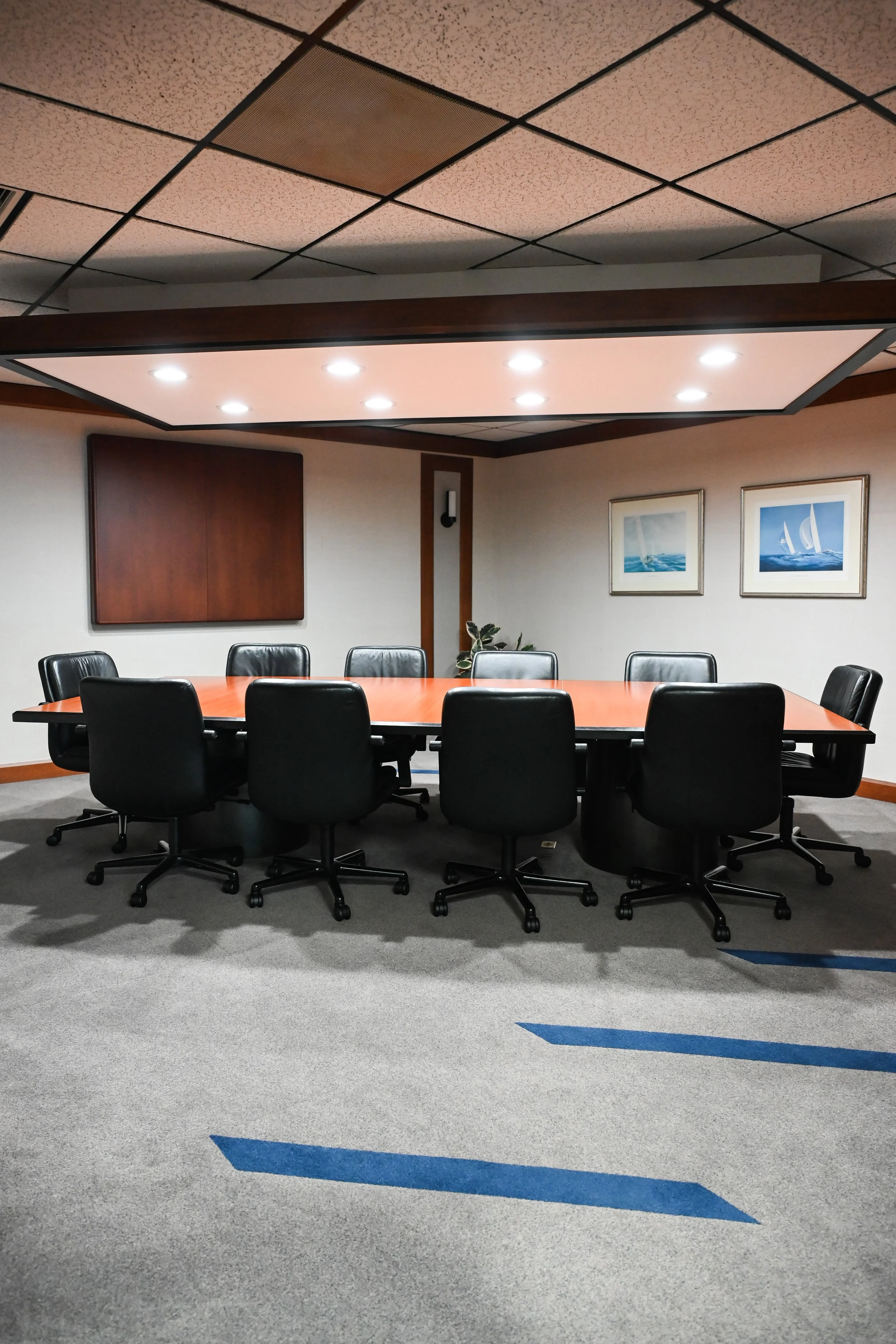 Empty conference room with ten black office chairs around a large wooden table, framed pictures of sailboats on the wall, a small plant, and ceiling lights.